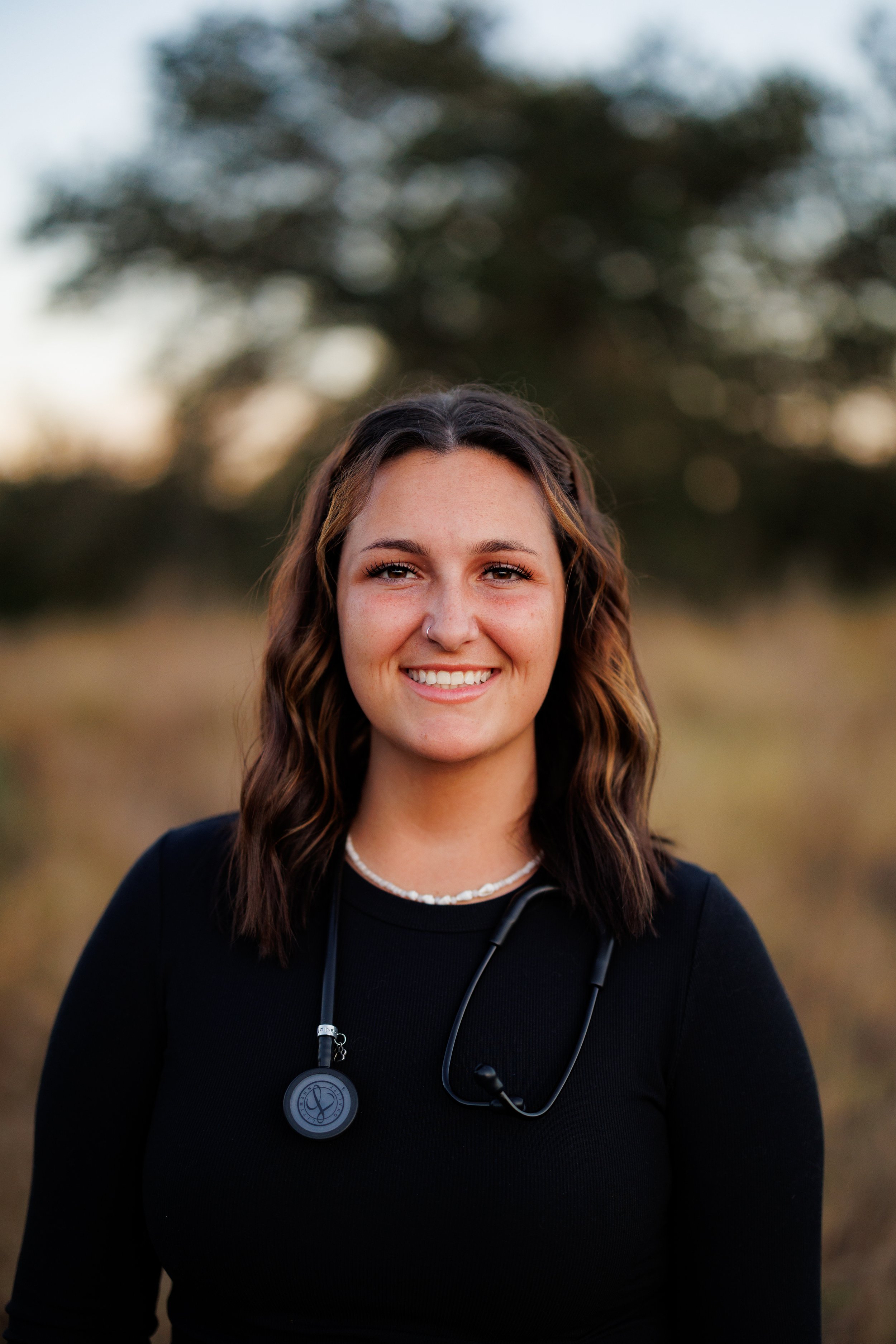 A young woman with shoulder-length brown hair, smiling, wearing a black top, with a stethoscope around her neck, standing outdoors during sunset.