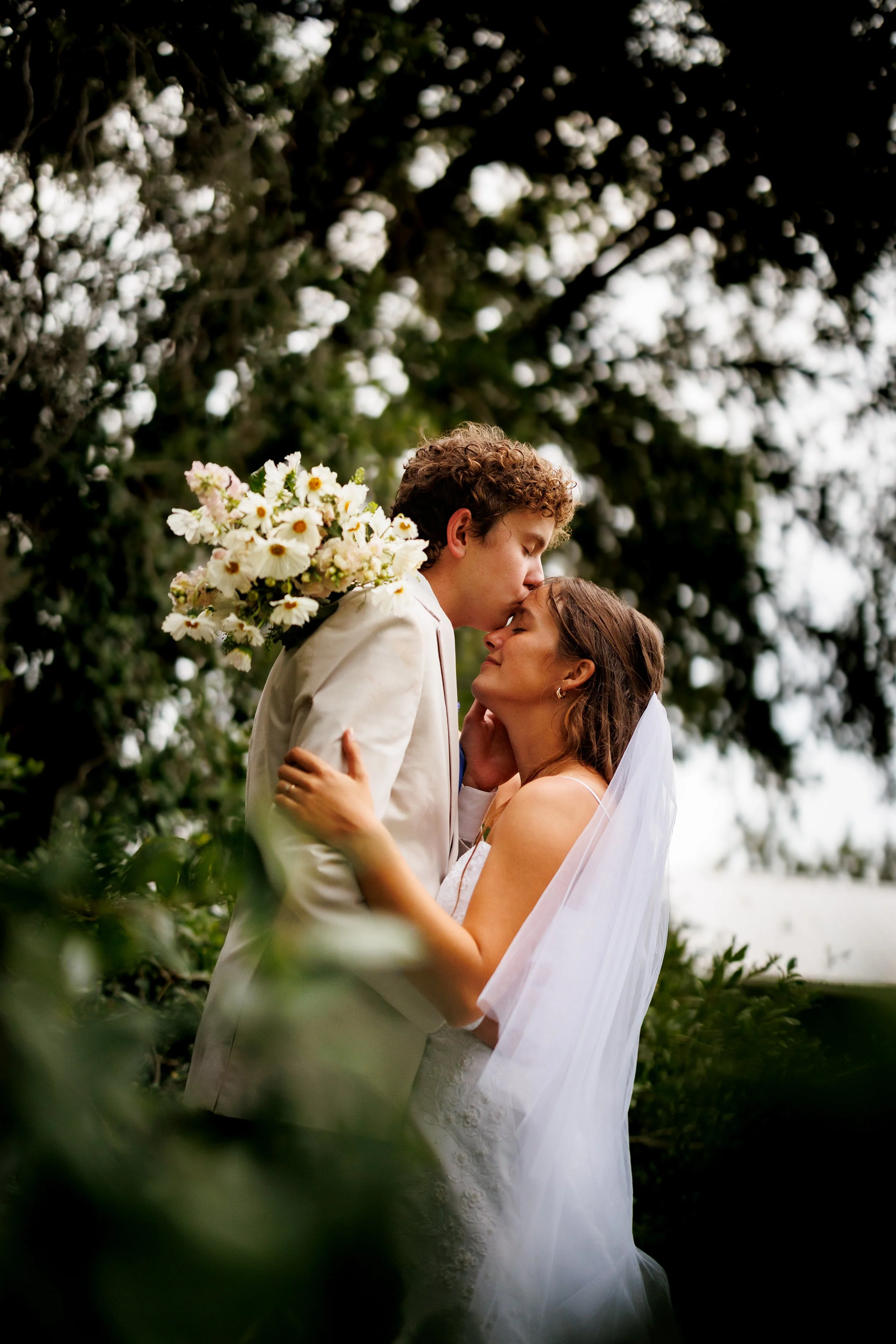 A bride and groom embrace outdoors, sharing a kiss. The groom holds a bouquet of white flowers over his shoulder. They are surrounded by greenery and trees, with the bride wearing a white wedding dress and veil, and the groom in a white suit.