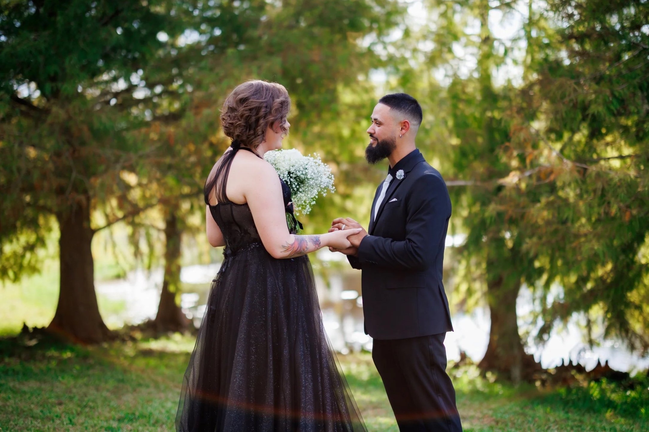 A couple standing outdoors exchanging vows, the woman in a black dress holding a bouquet, and the man in a black suit, with trees and sunlight in the background.