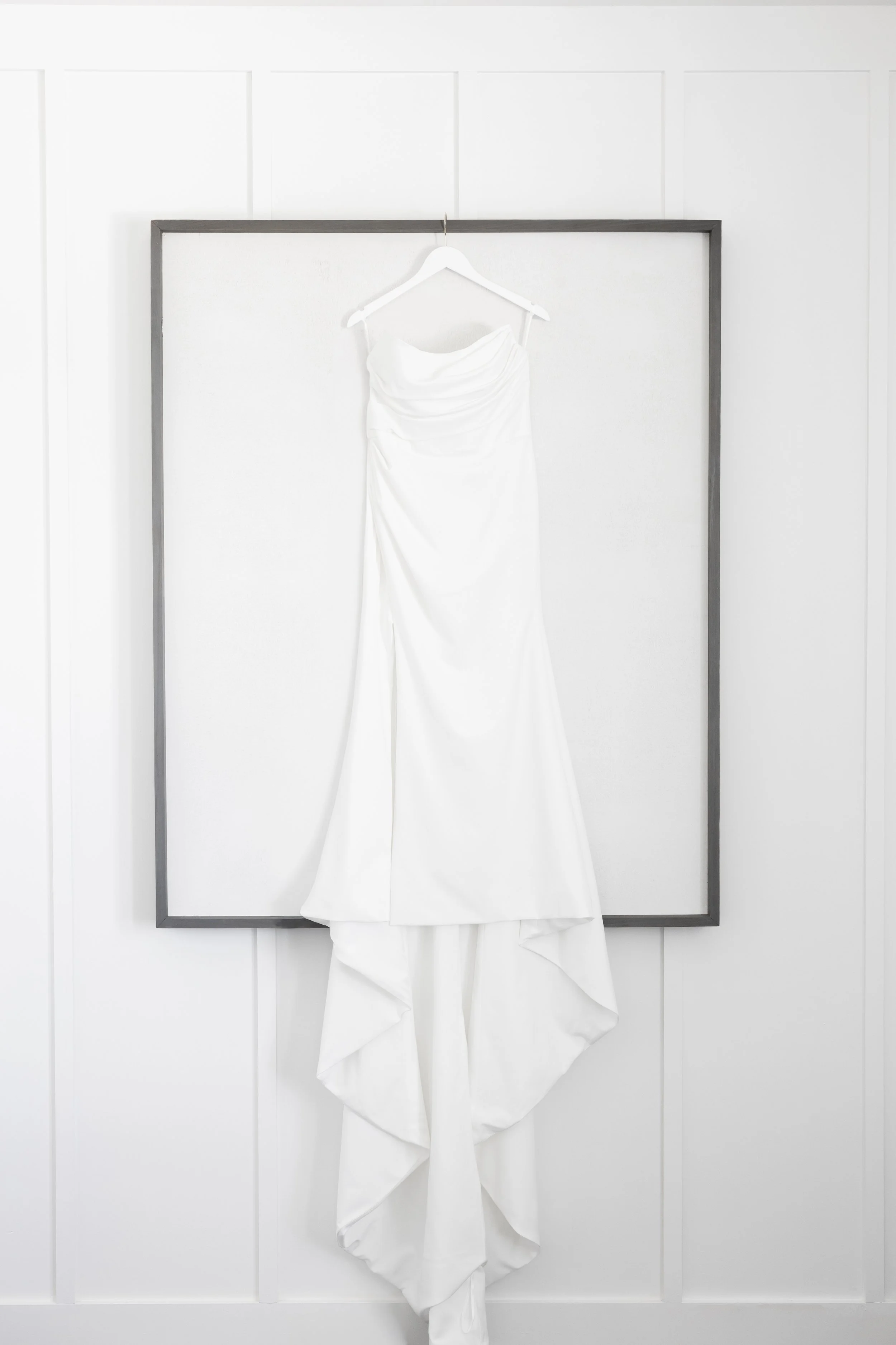 White wedding dress hanging on a white hanger against a white wall framed by a dark gray rectangle.