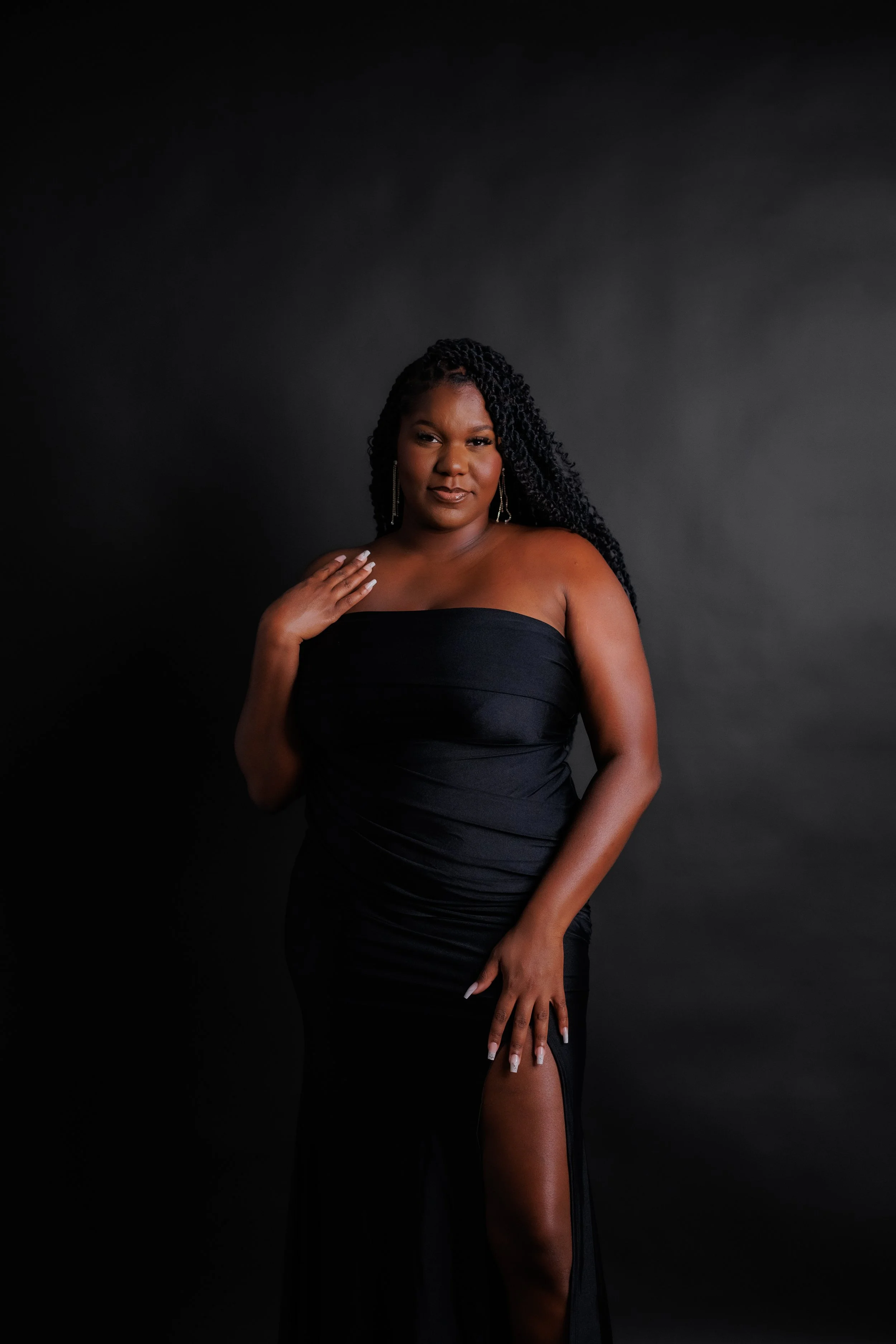 A woman with dark, wavy braided hair wearing a strapless black gown with a high slit, standing against a dark background.