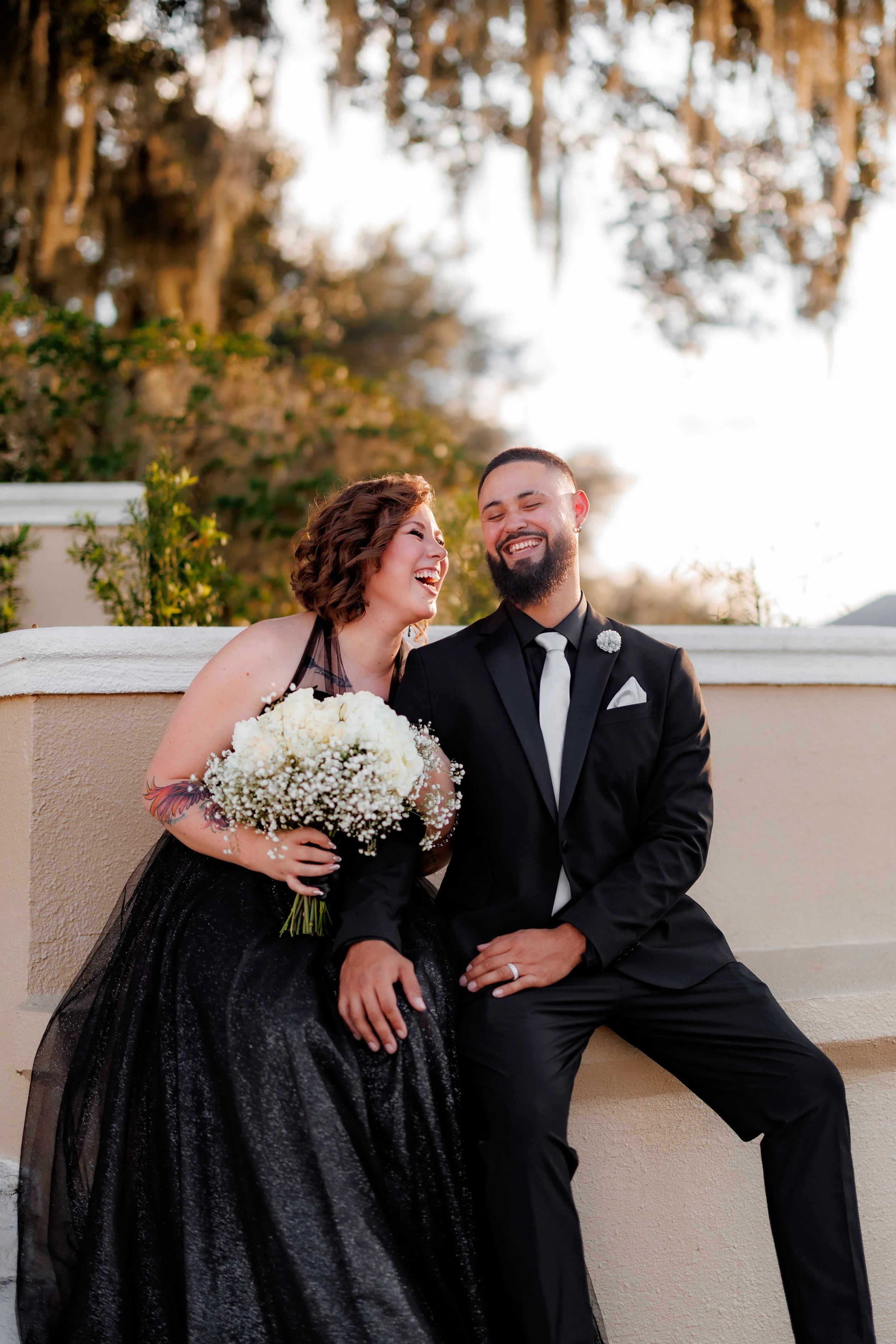 A newlywed couple sitting on a wall, smiling and laughing together outside during sunset, with trees in the background. The bride is holding a bouquet of white flowers and wearing a black dress, while the groom is dressed in a black suit with a white