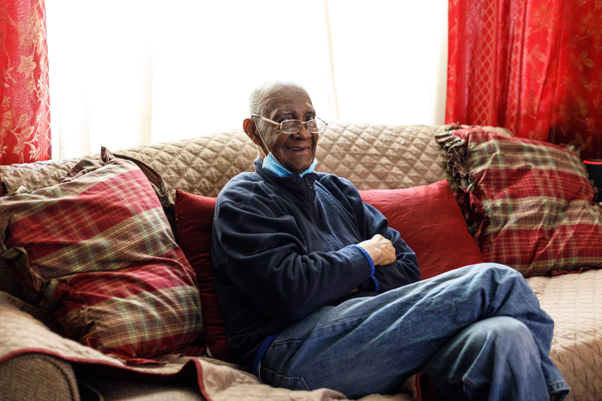An elderly man sitting on a couch with several plaid and solid red pillows, smiling, wearing glasses and a face mask pulled down, in a room with red curtains and natural light.