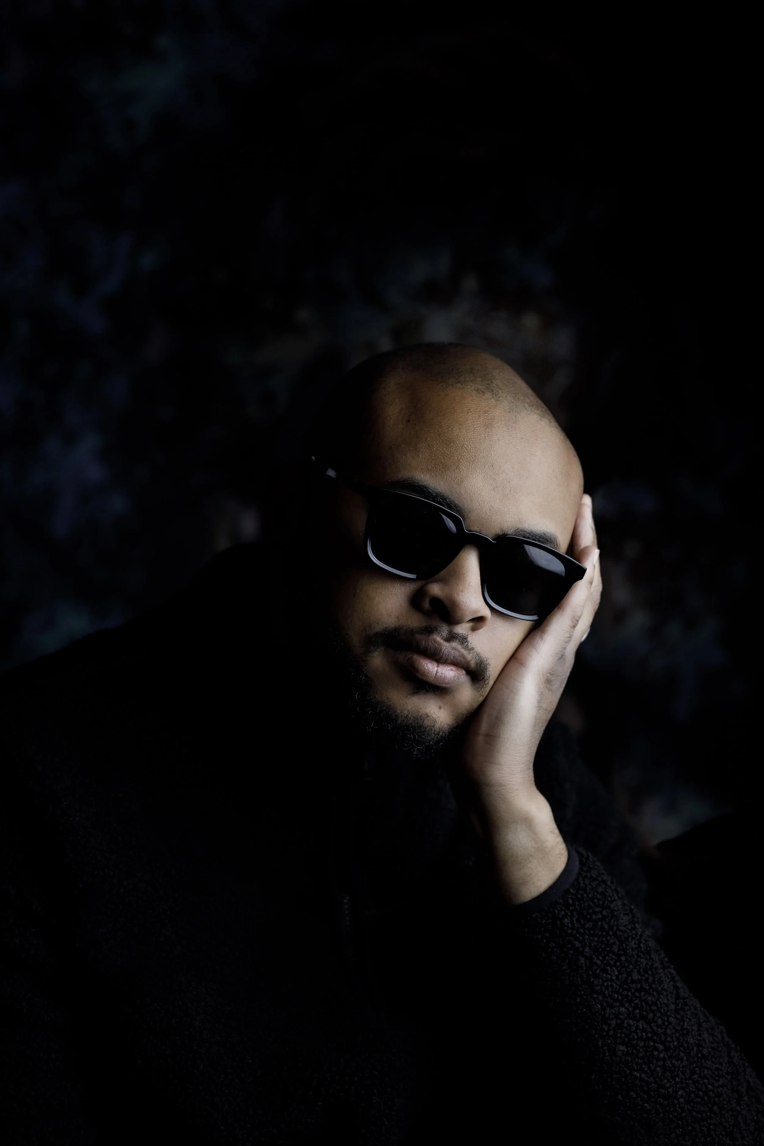 Portrait of a man with sunglasses, bald head, and beard, resting his face on his hand against a dark background.