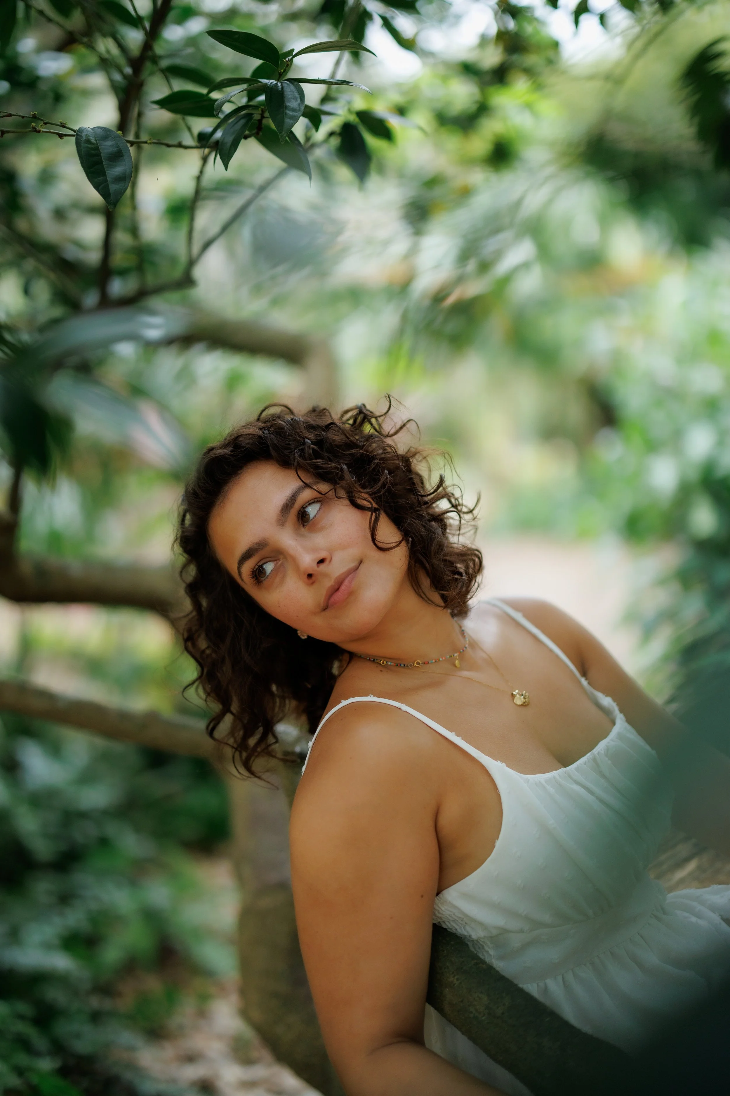 A young woman with curly hair wearing a white dress and colorful necklaces, leaning on a wooden railing in a lush green garden.