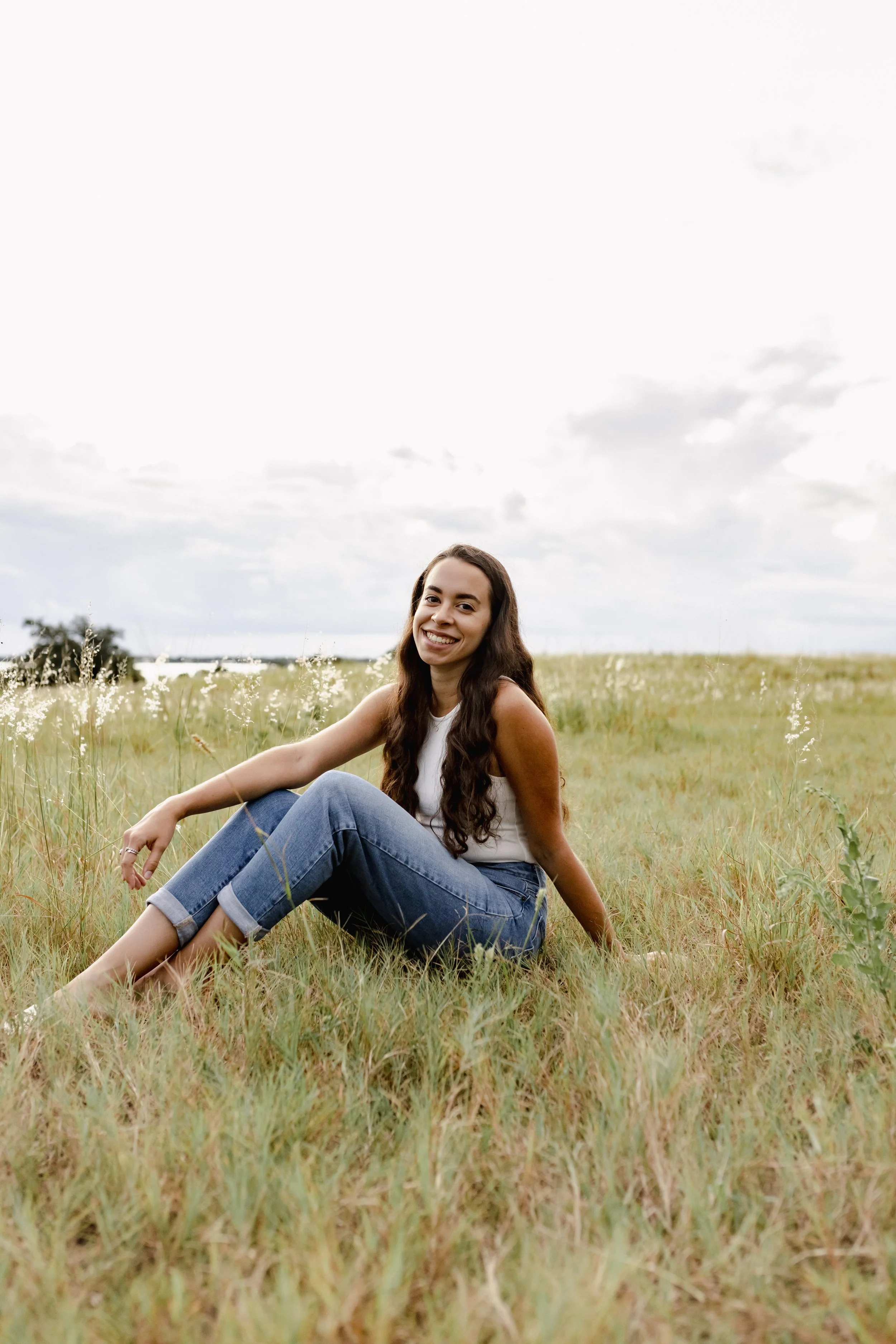 A young woman with long dark hair, smiling, sitting on a grassy field during daytime, wearing a white sleeveless top and rolled-up jeans.