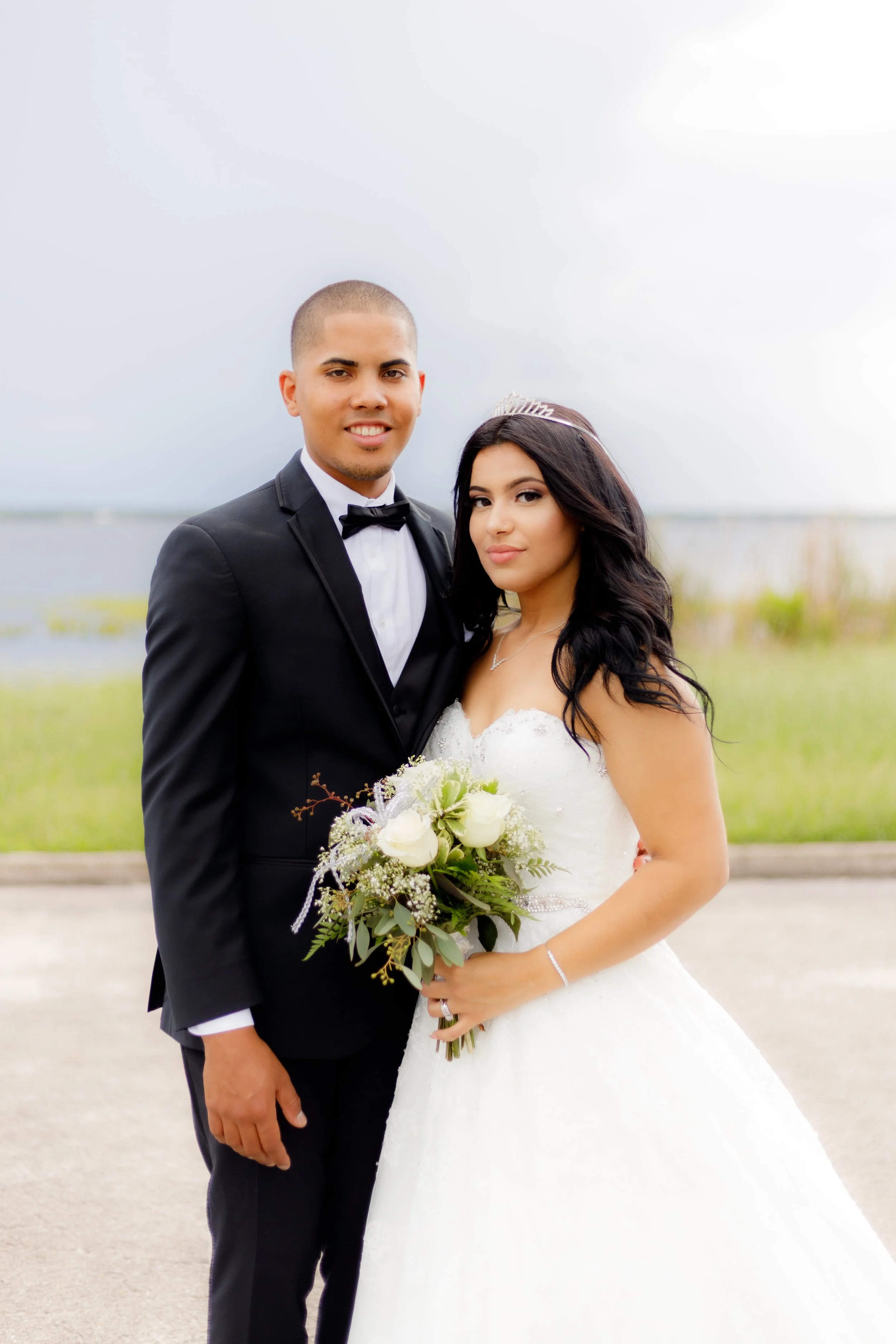 A newlywed couple standing outdoors by a body of water, dressed in wedding attire with the man in a black tuxedo and the woman in a white wedding gown holding a bouquet of white roses.