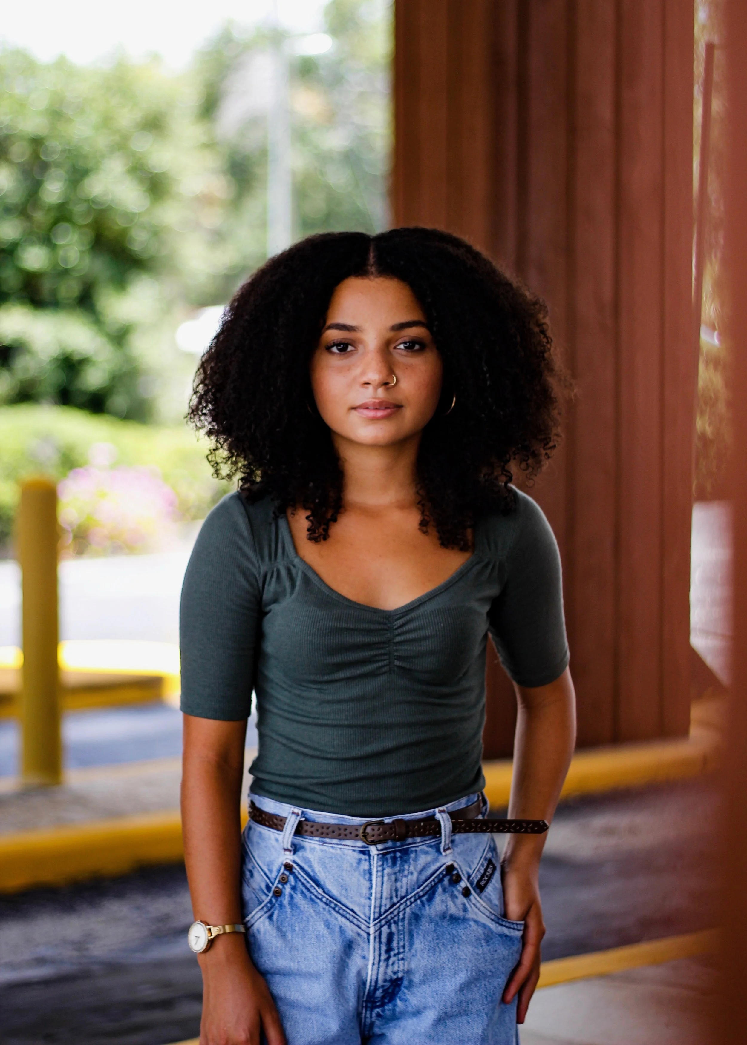 A young woman with curly black hair, wearing a dark green top, jeans, and a watch, standing outside near a wooden wall and greenery, looking at the camera.