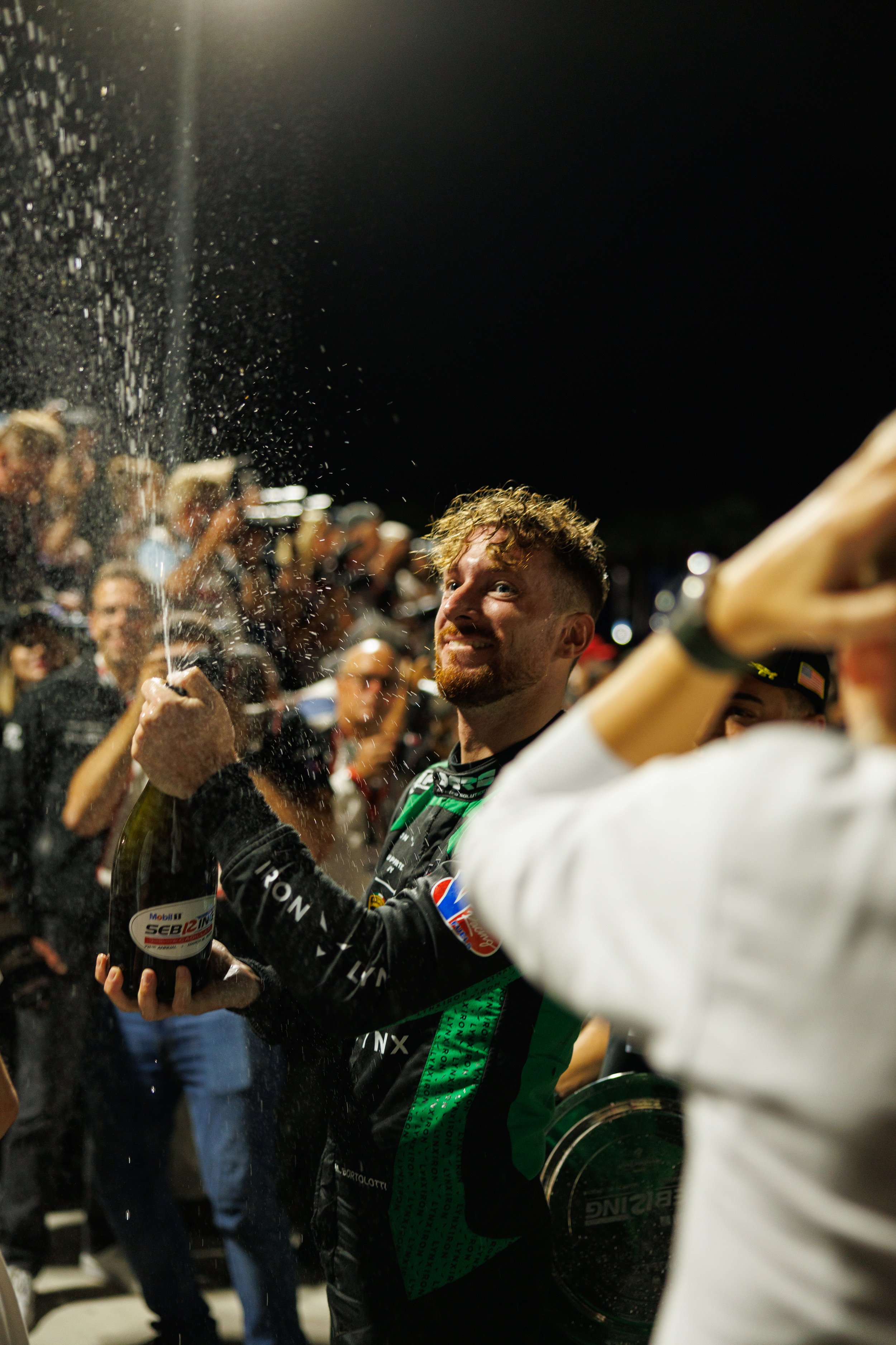 A race car driver celebrating in victory lane, holding a bottle of champagne, with a spray of champagne in the air, surrounded by fans and team members at night.