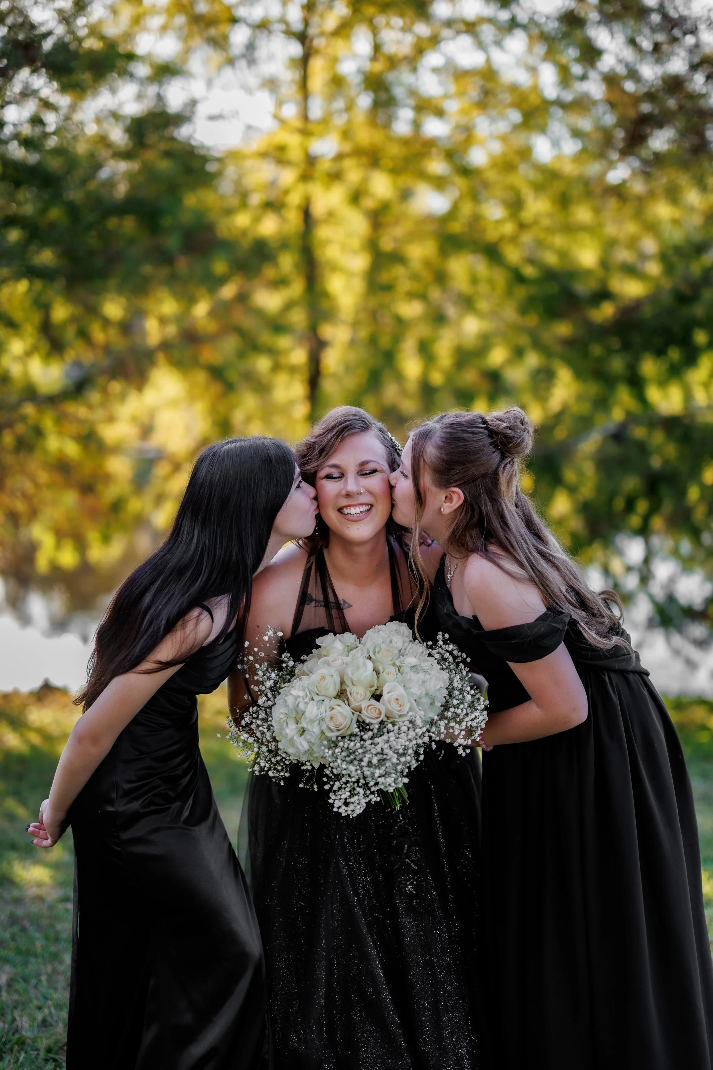 A bride in a black dress holding a bouquet of white roses is kissing two women, also in black dresses, on the cheeks during a wedding celebration outdoors with trees in the background.