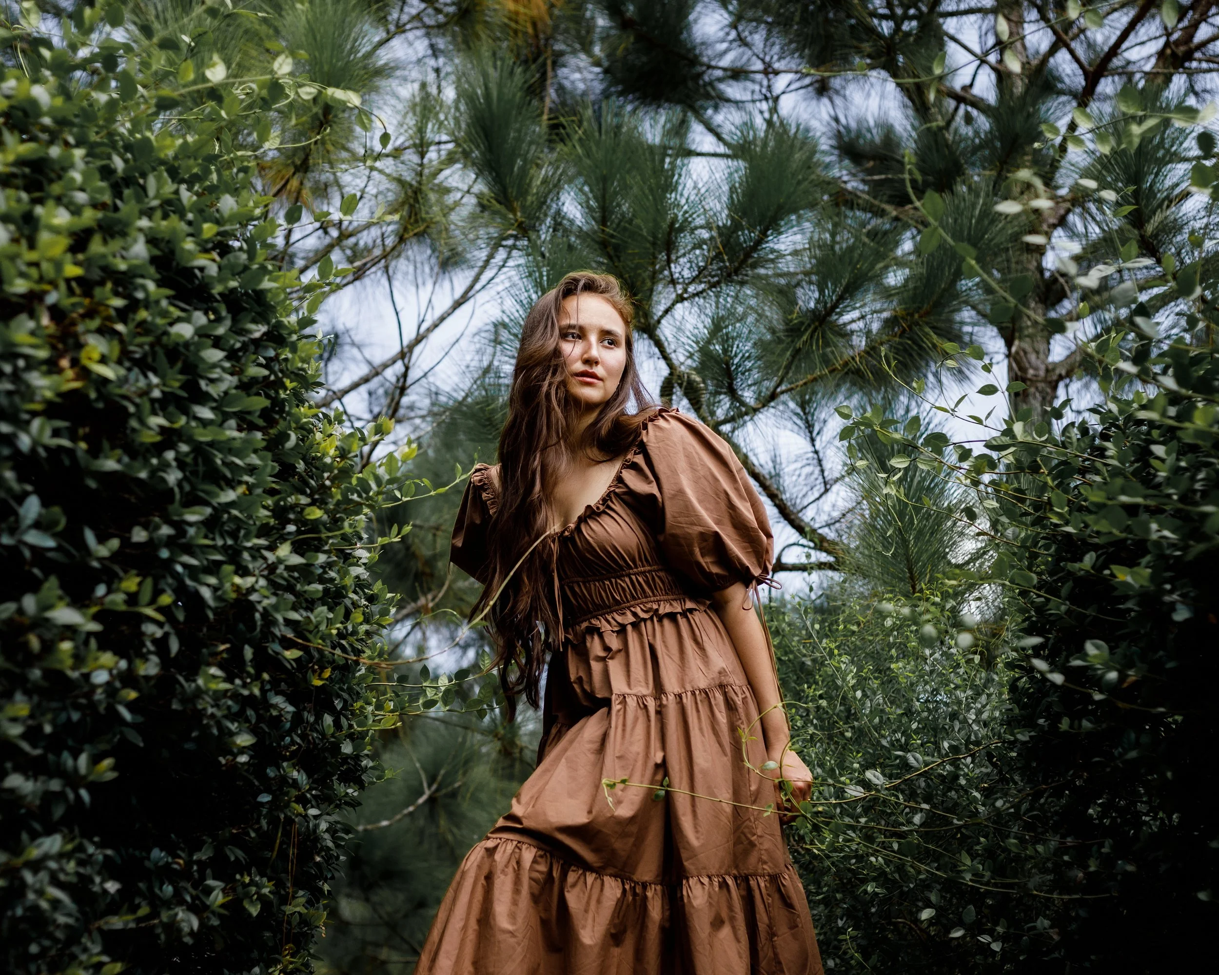 A young woman with long wavy brown hair standing among green foliage and trees outdoors, wearing a brown dress with puffed sleeves and tiered skirt, looking slightly to her right.