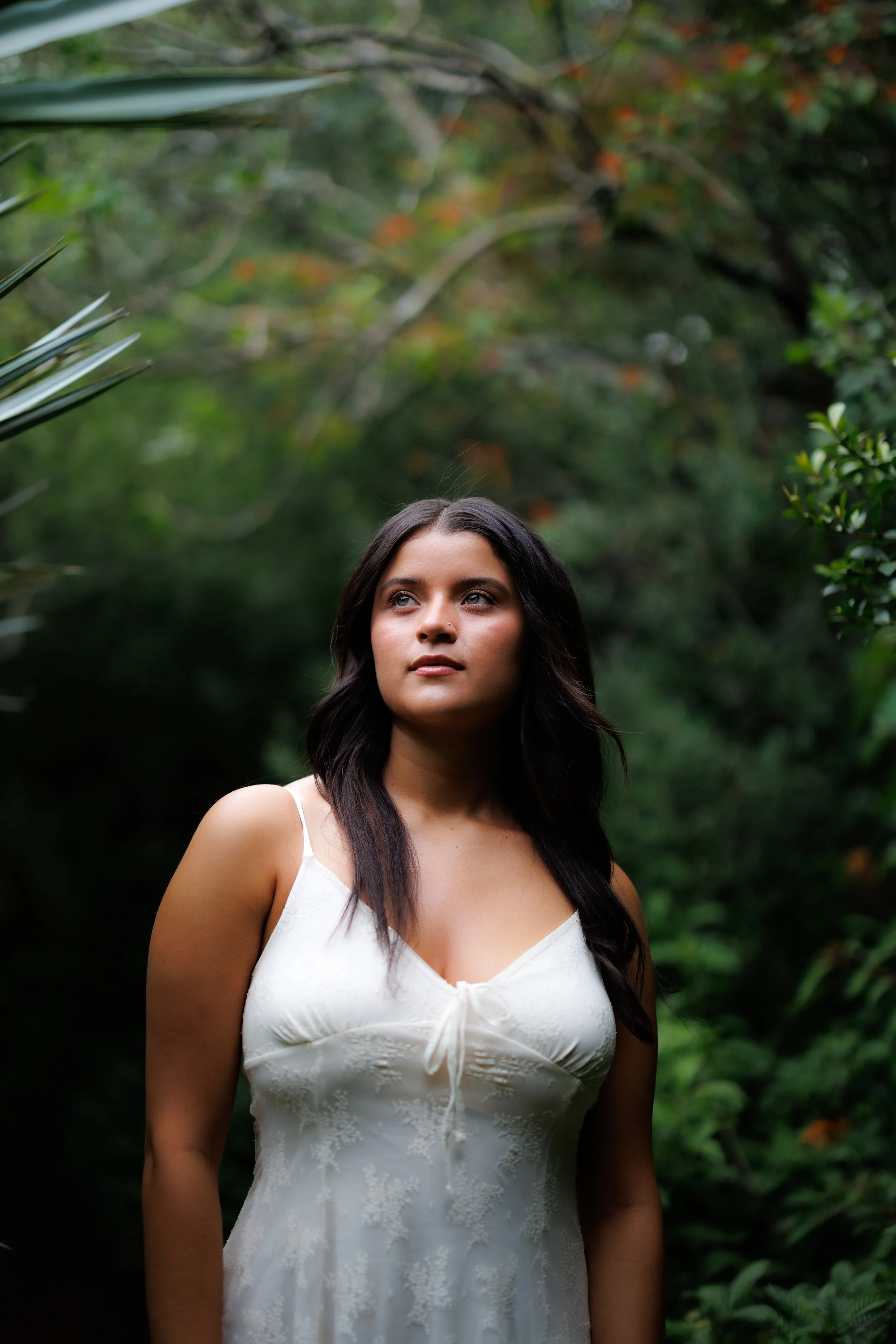 A woman with dark hair wearing a white dress standing amidst lush green foliage in a forest or garden setting.