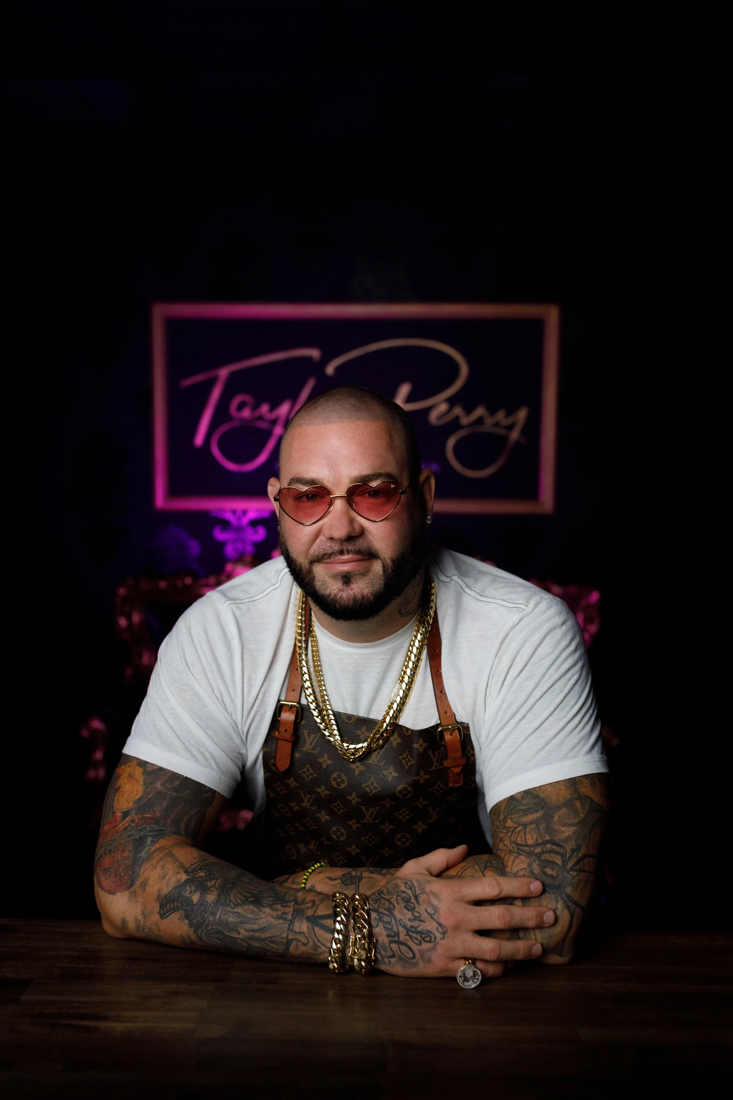 A man with tattoos, wearing sunglasses and gold chains, sitting at a table with a dark background and a sign that says "Taylour Perry" with pink and purple lighting.