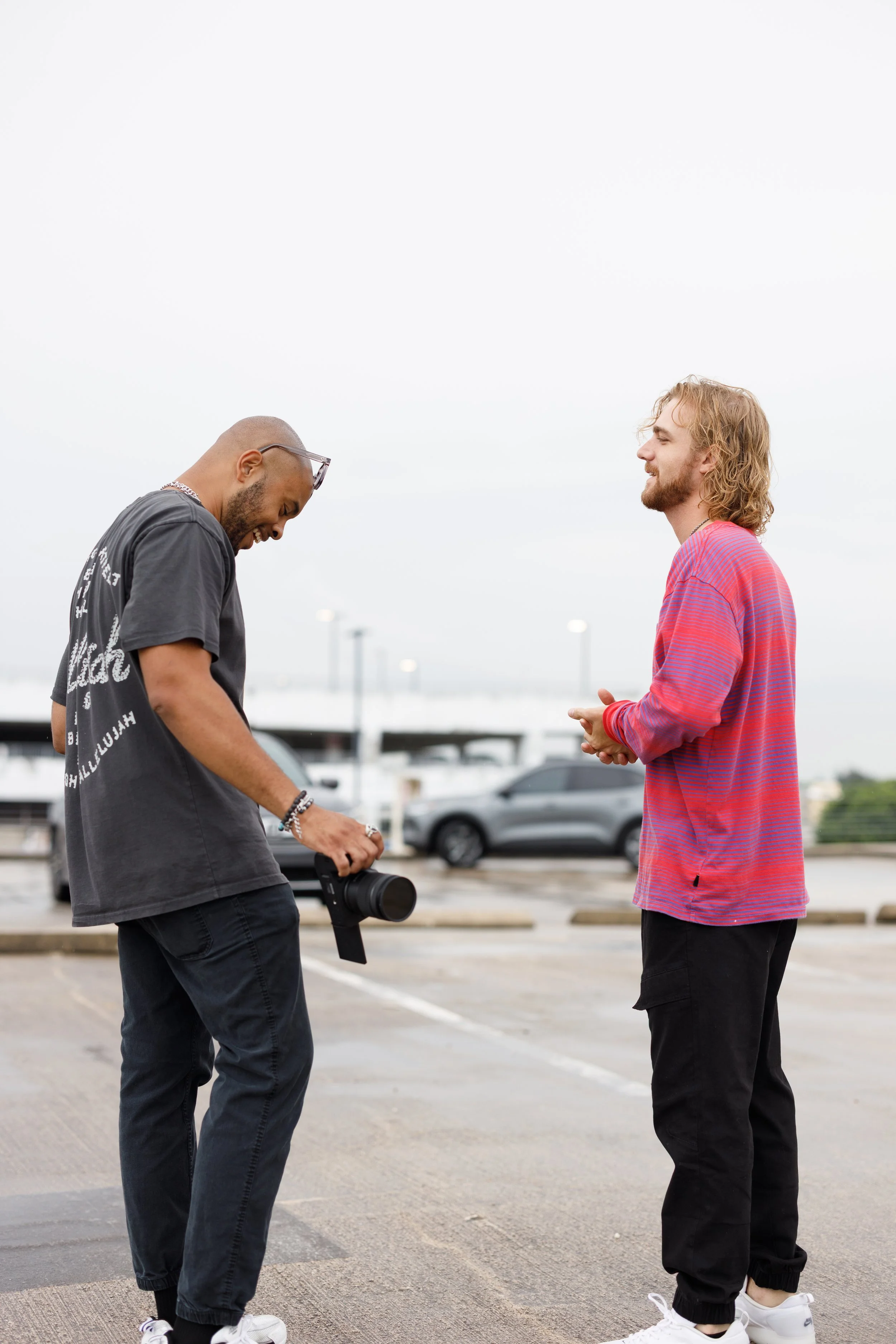 Two men standing in a parking lot, one holding a camera and the other smiling, engaged in conversation on an overcast day.