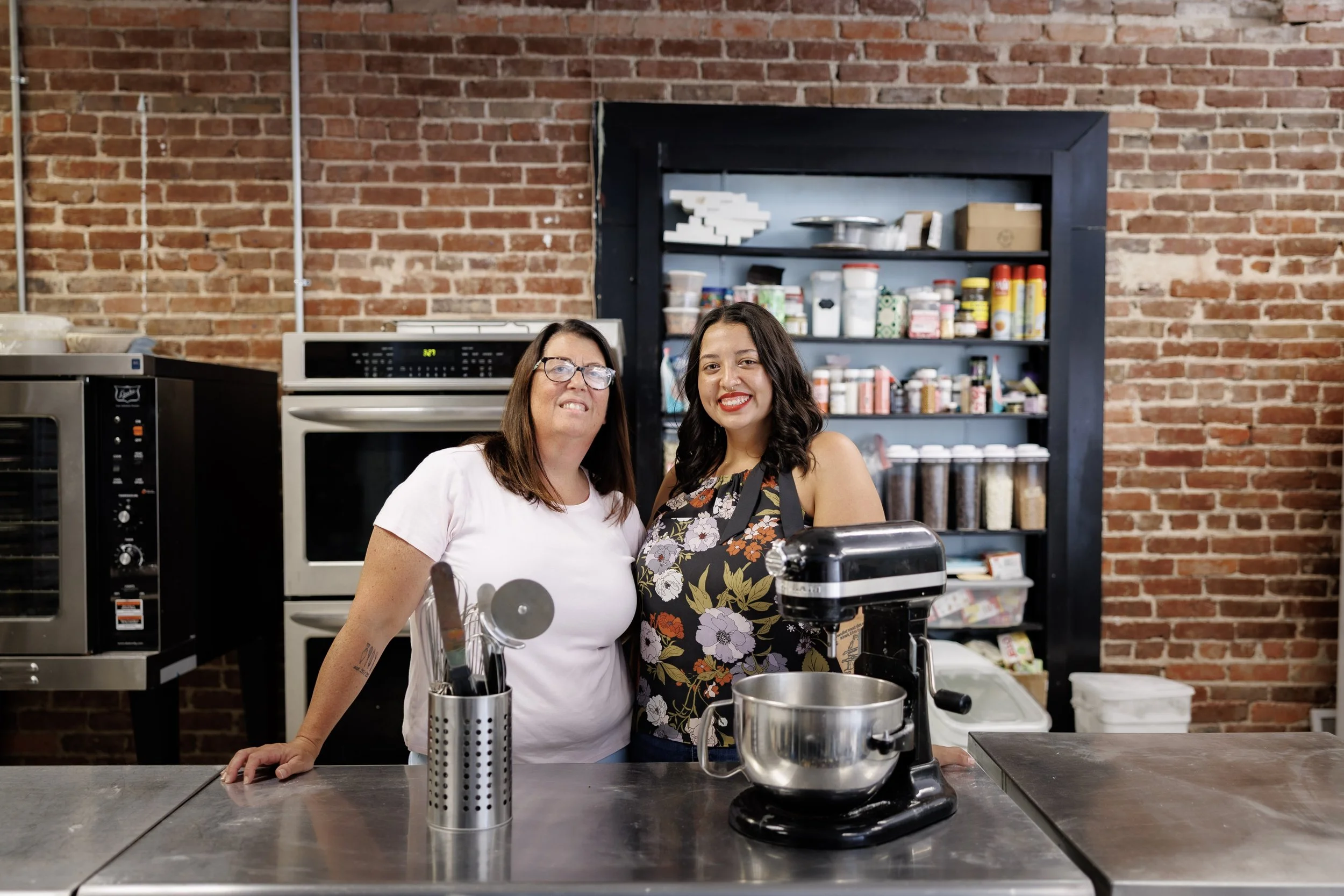 Two women standing behind a kitchen counter in a cafe, smiling at the camera, with a brick wall and shelf with kitchen supplies in the background.