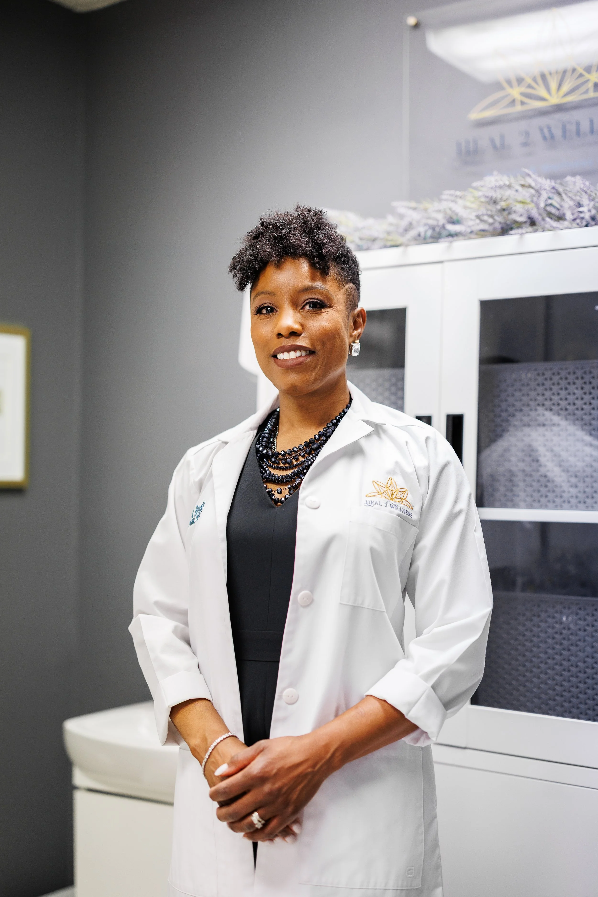 A woman in a white lab coat standing in a medical or wellness clinic, smiling at the camera, wearing a black dress and layered black necklaces, with a cabinet and lavender flowers in the background.