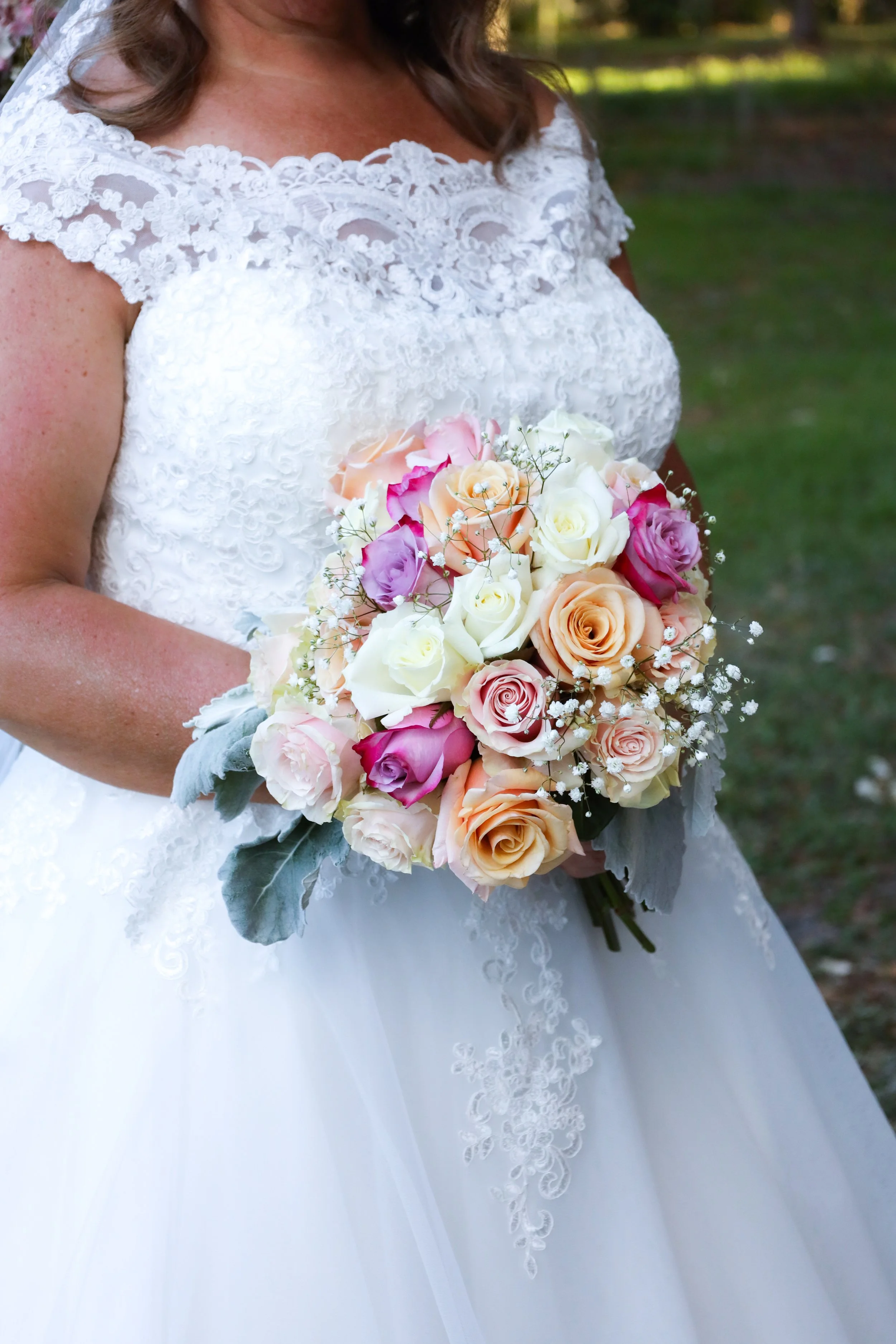 Bride in a white lace wedding dress holding a colorful bouquet of roses and baby's breath, standing outdoors.