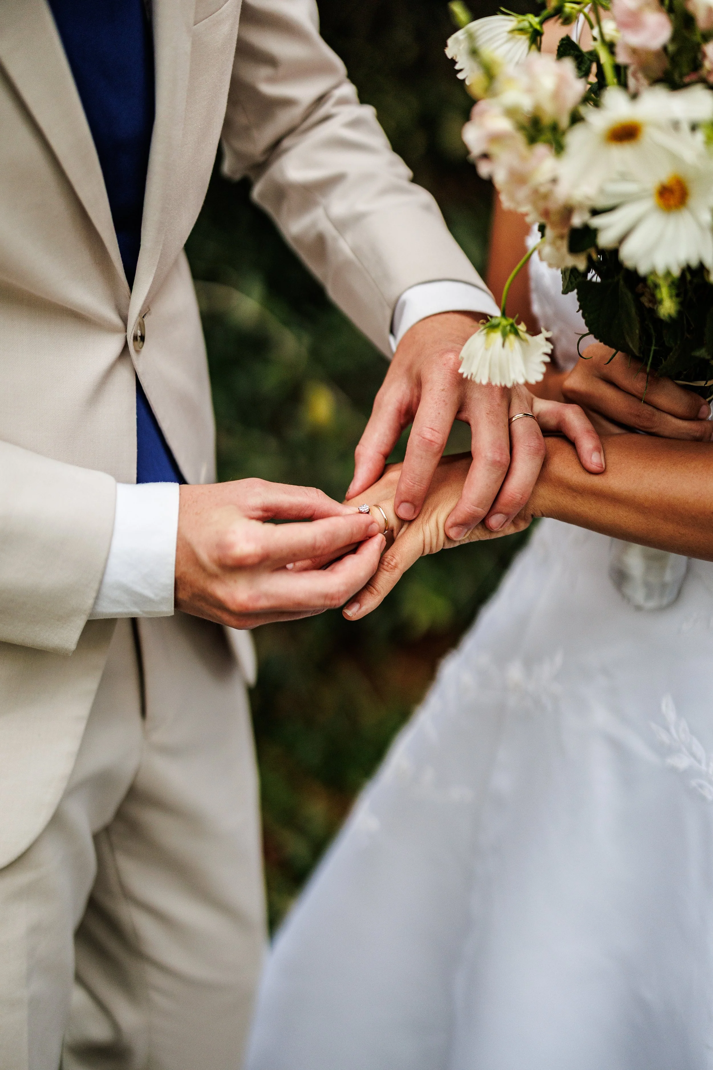 A couple is getting married, with one person placing a ring on the other's finger, holding her hand and a bouquet of flowers in the background.