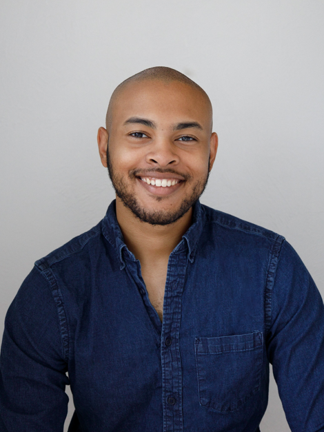 Portrait of a smiling man with a beard, wearing a dark blue button-up shirt, against a plain light gray background.