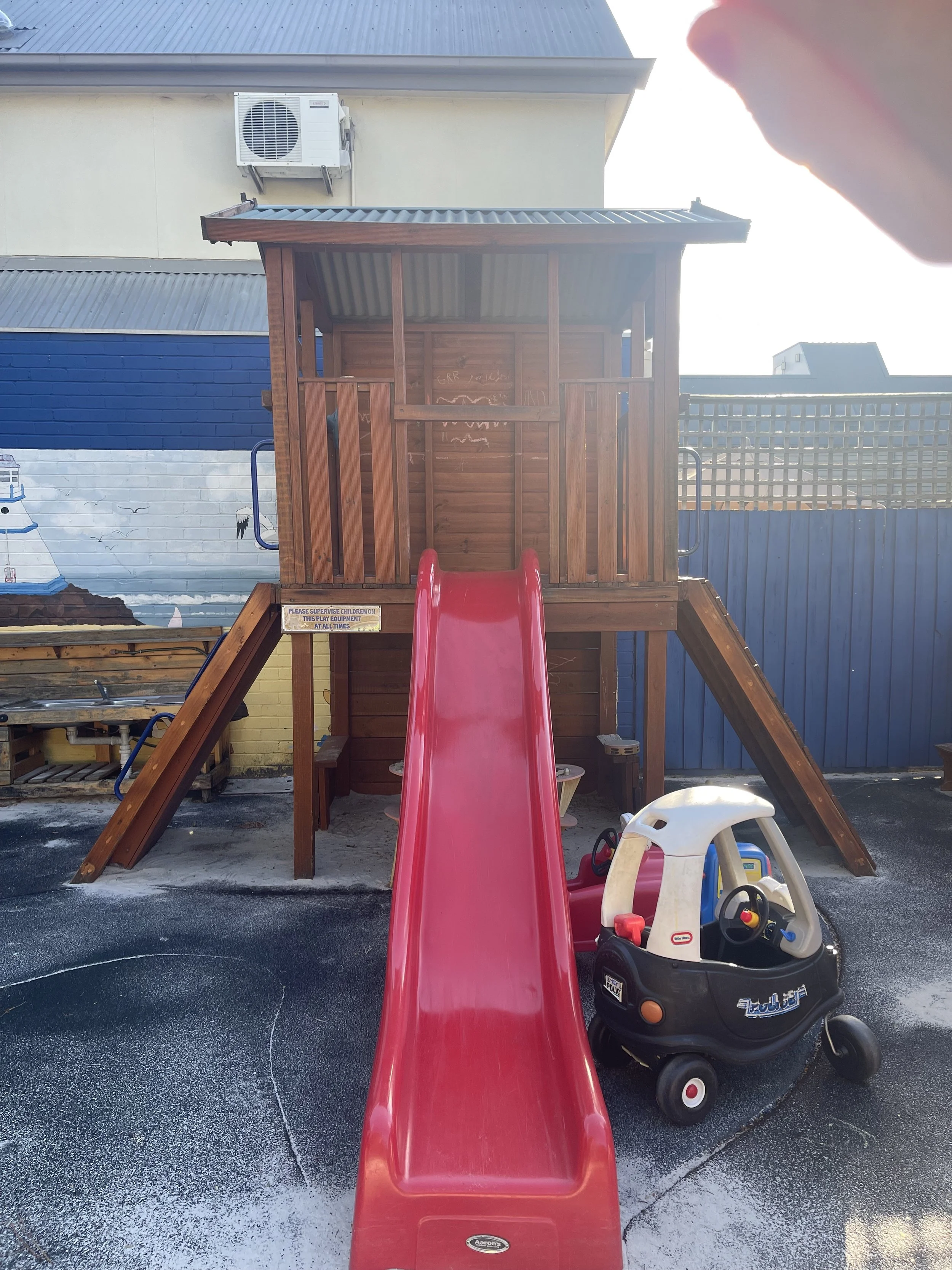 A children's wooden playhouse with a red slide and a small toy car in front, located outdoors on a blacktop surface.