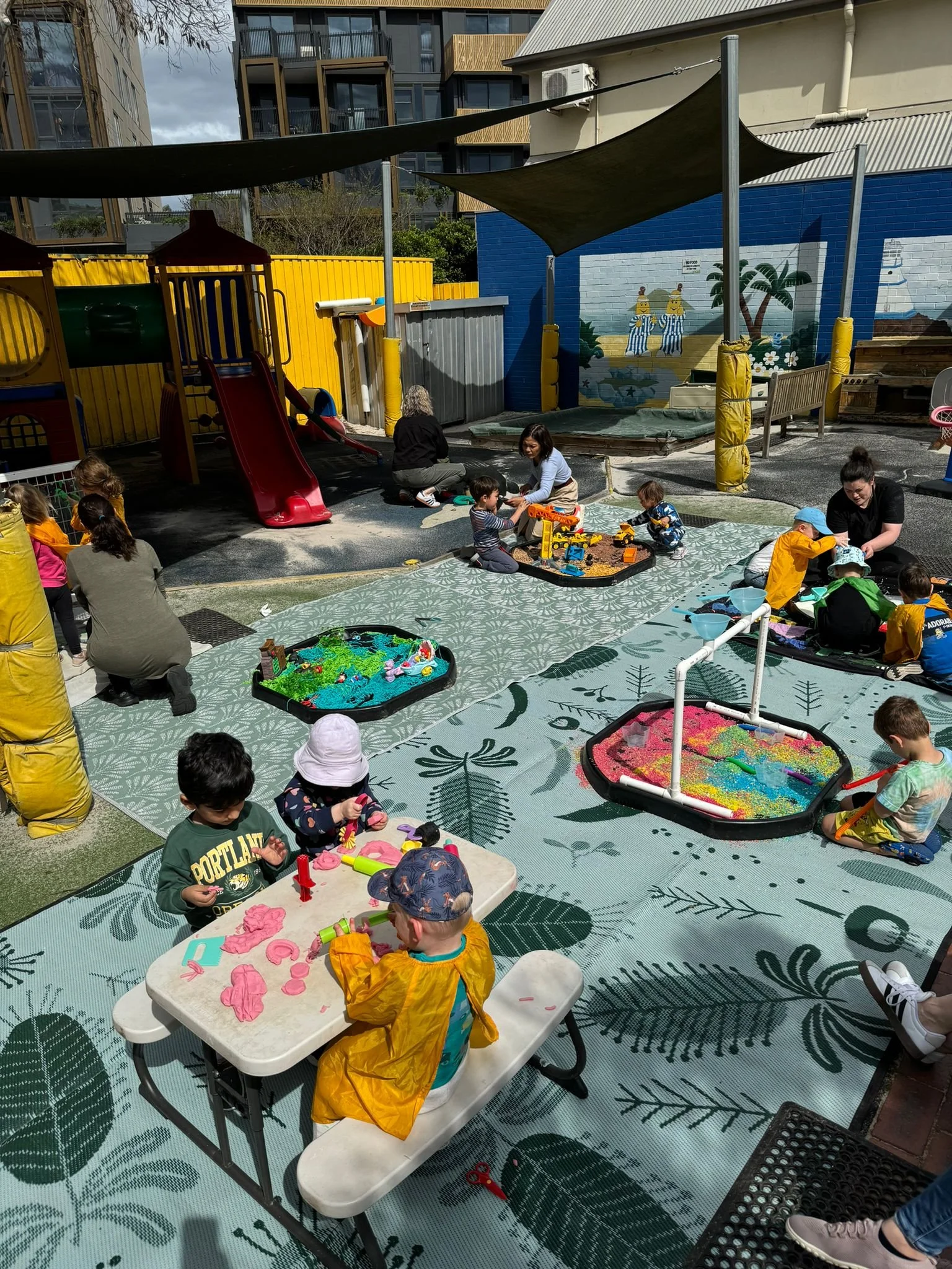 Children and adults at an outdoor playground and sensory activity area with pools of water, sand, and craft activities, surrounded by colorful play structures and shaded canopy.