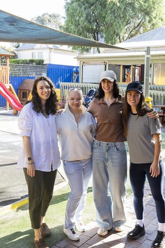 Four women smiling and posing together outdoors in a playground area with a slide and fencing in the background.