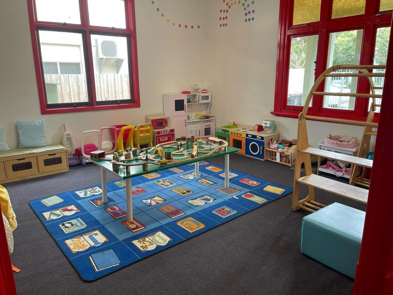Children's playroom with a train set on a table, a colorful rug with alphabet and animal images, toy kitchen set, doll furniture, and large windows with red frames.