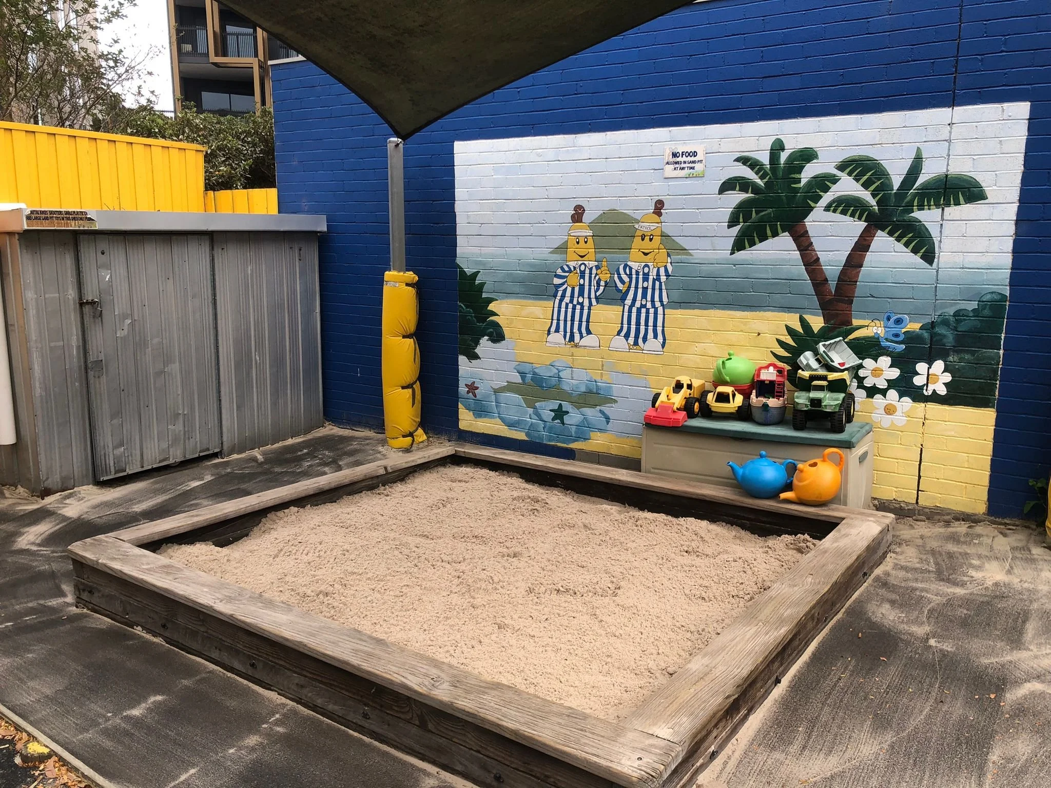 A colorful children's play area with a sandpit surrounded by a wooden border, a mural of cartoon characters and tropical trees painted on the wall, and various plastic toys including trucks, teapots, and watering cans.