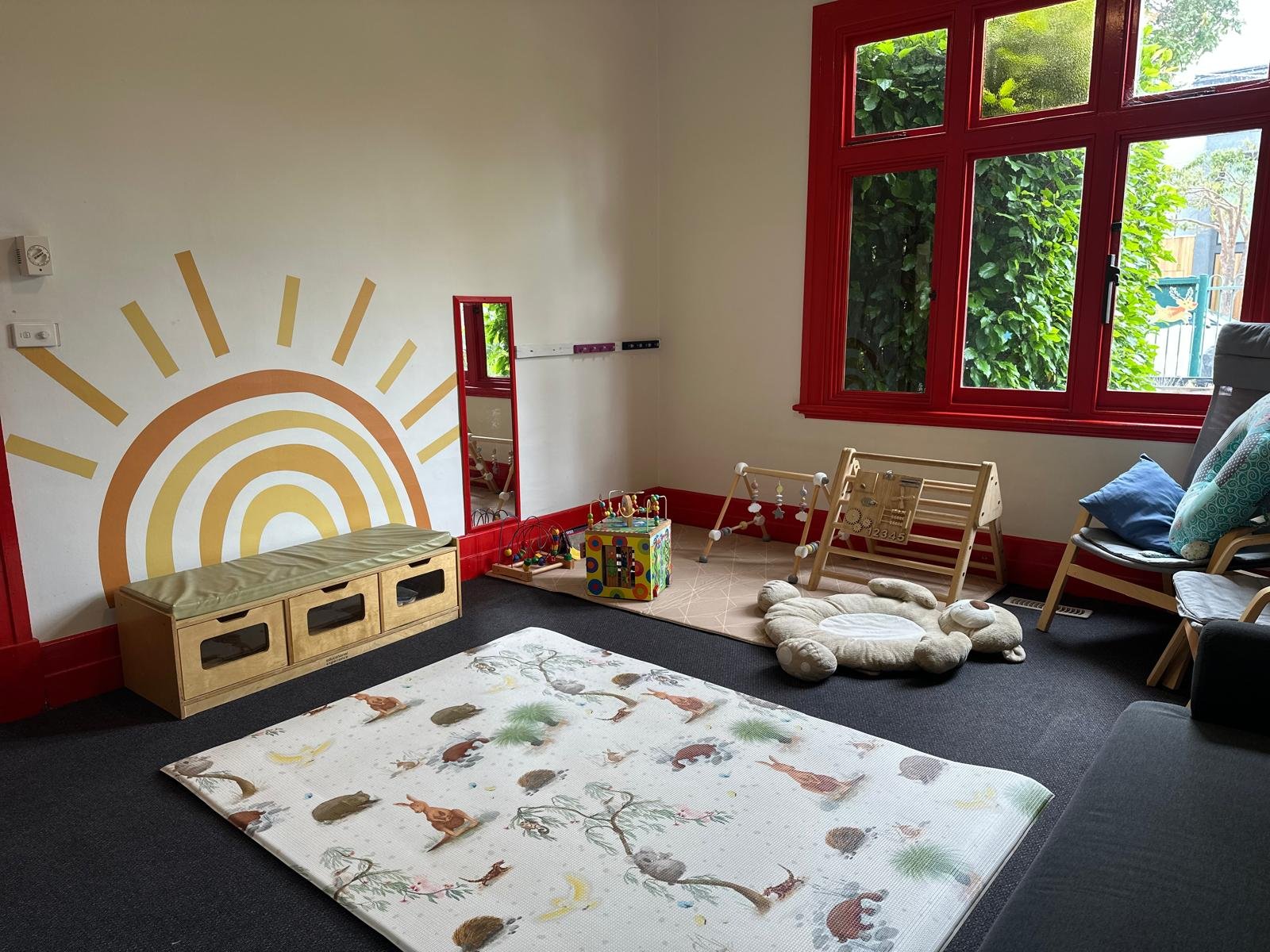 Child's playroom with a sun mural on the wall, window with red frame, children's toys, a rug with animal illustrations, a stuffed animal seat, a small chair with pillows, and various toys and furniture.
