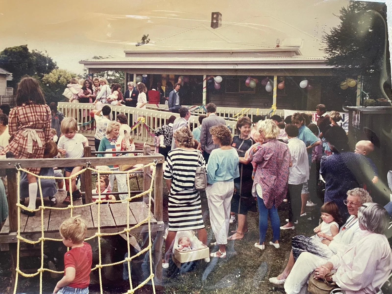 A large gathering of children and adults at a backyard party, with some children playing on a wooden playground and others standing or sitting around, near a house decorated with balloons and streamers.