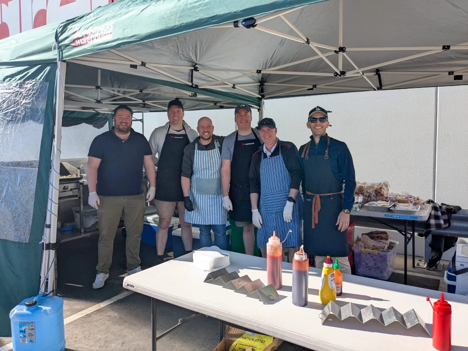 Group of seven men standing under a canopy tent, smiling, with grilling and condiments on a table in front of them, at an outdoor event.