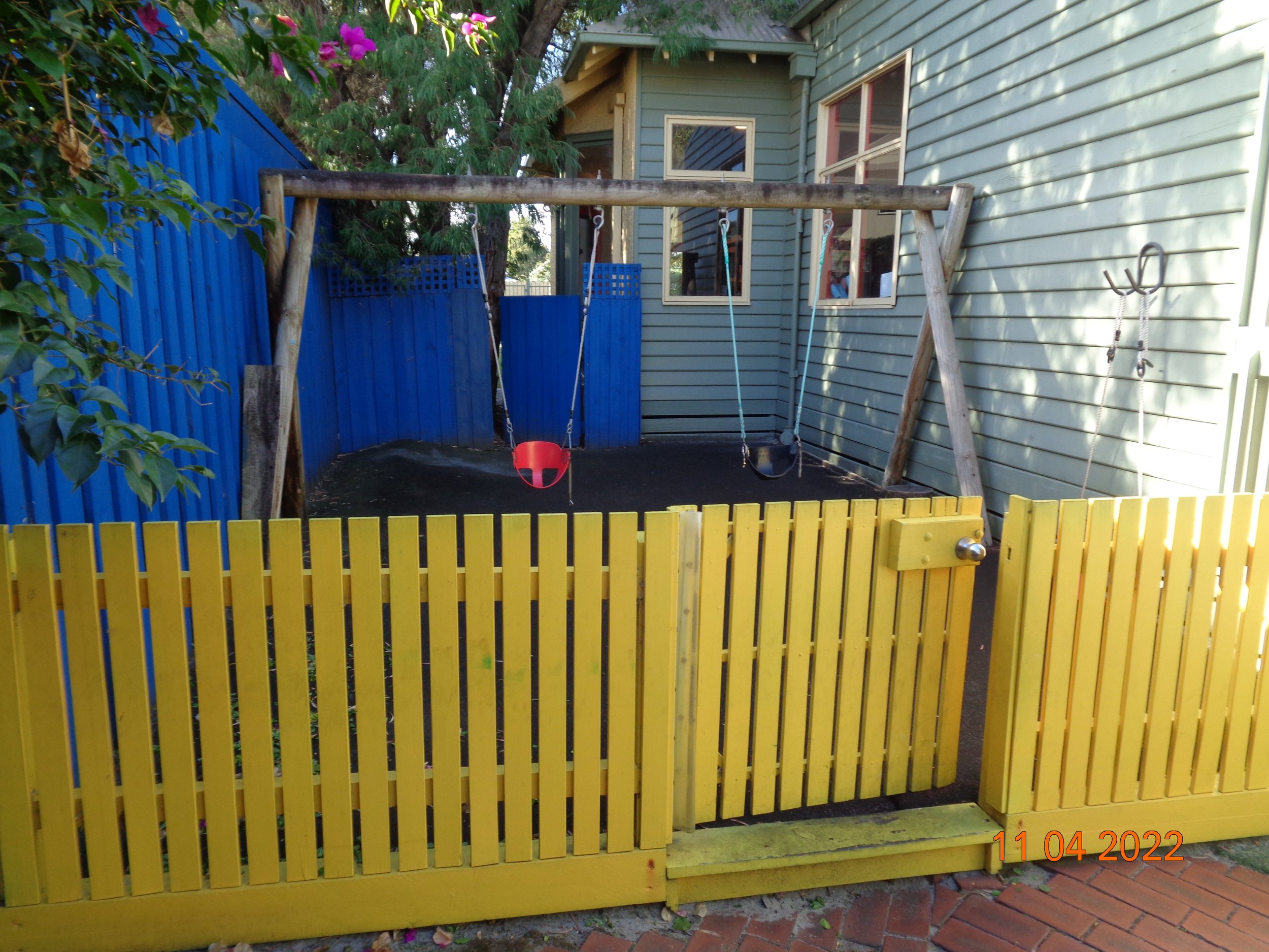 A small backyard with a wooden swing set that has two swings, one with a red seat and the other empty. The yard is enclosed by a bright yellow wooden fence and a blue fence in the background. The ground is covered with black rubber matting. 