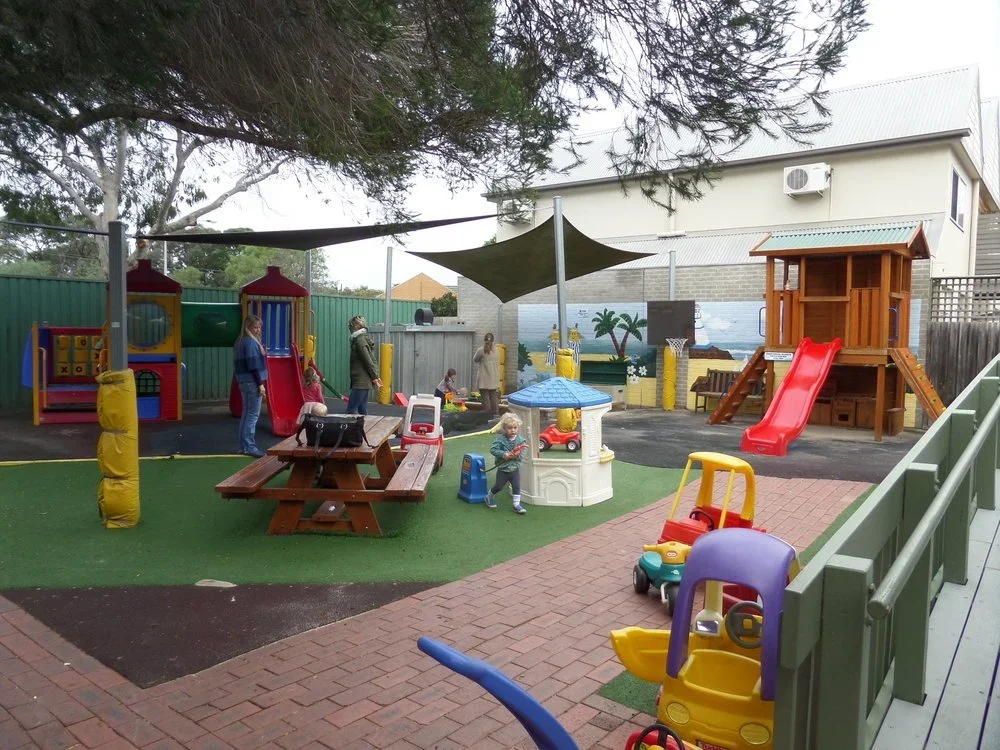 Children playing on playground equipment, including slides and a playhouse, with adults nearby, in a fenced area on a partly cloudy day.