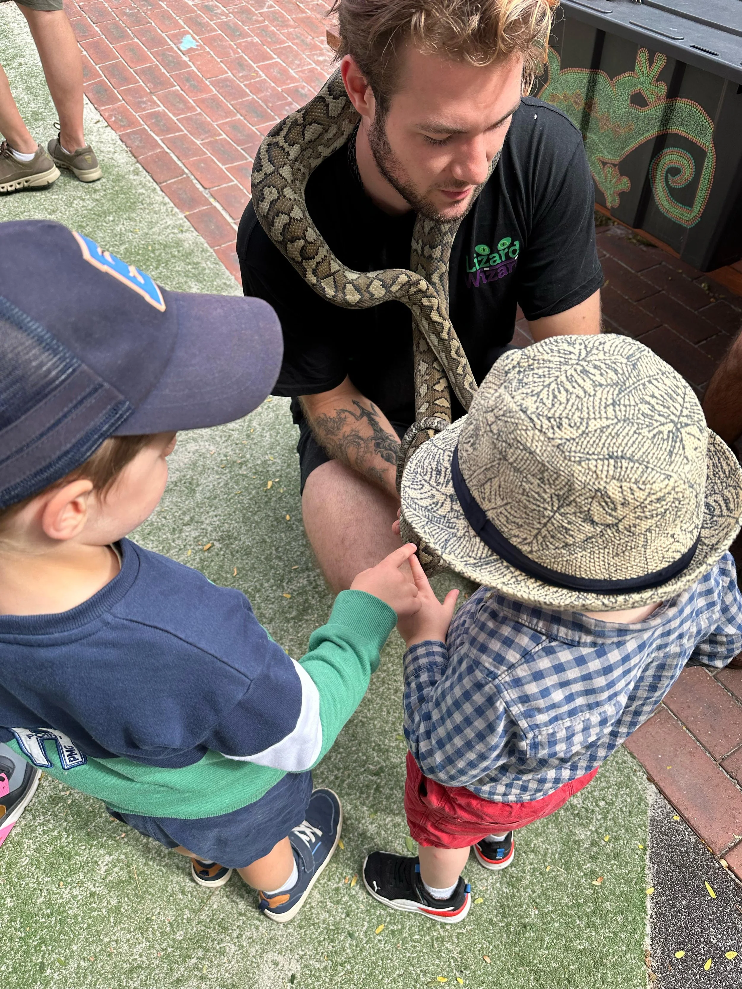 A man with a snake around his neck, interacting with two young children outdoors on a mixed surface of grass and brick.
