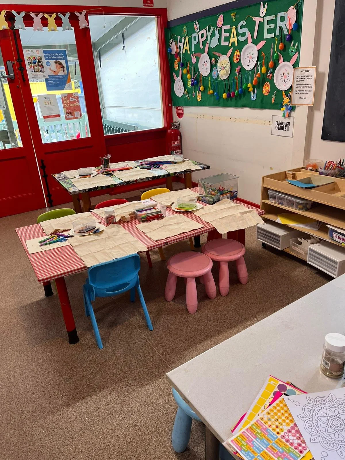 Easter themed classroom with a green bulletin board decorated with Easter eggs, bunnies, and the words 'Happy Easter'. There are tables with craft supplies, paper, markers, and scissors set up for children to make Easter crafts. Brightly colored chai
