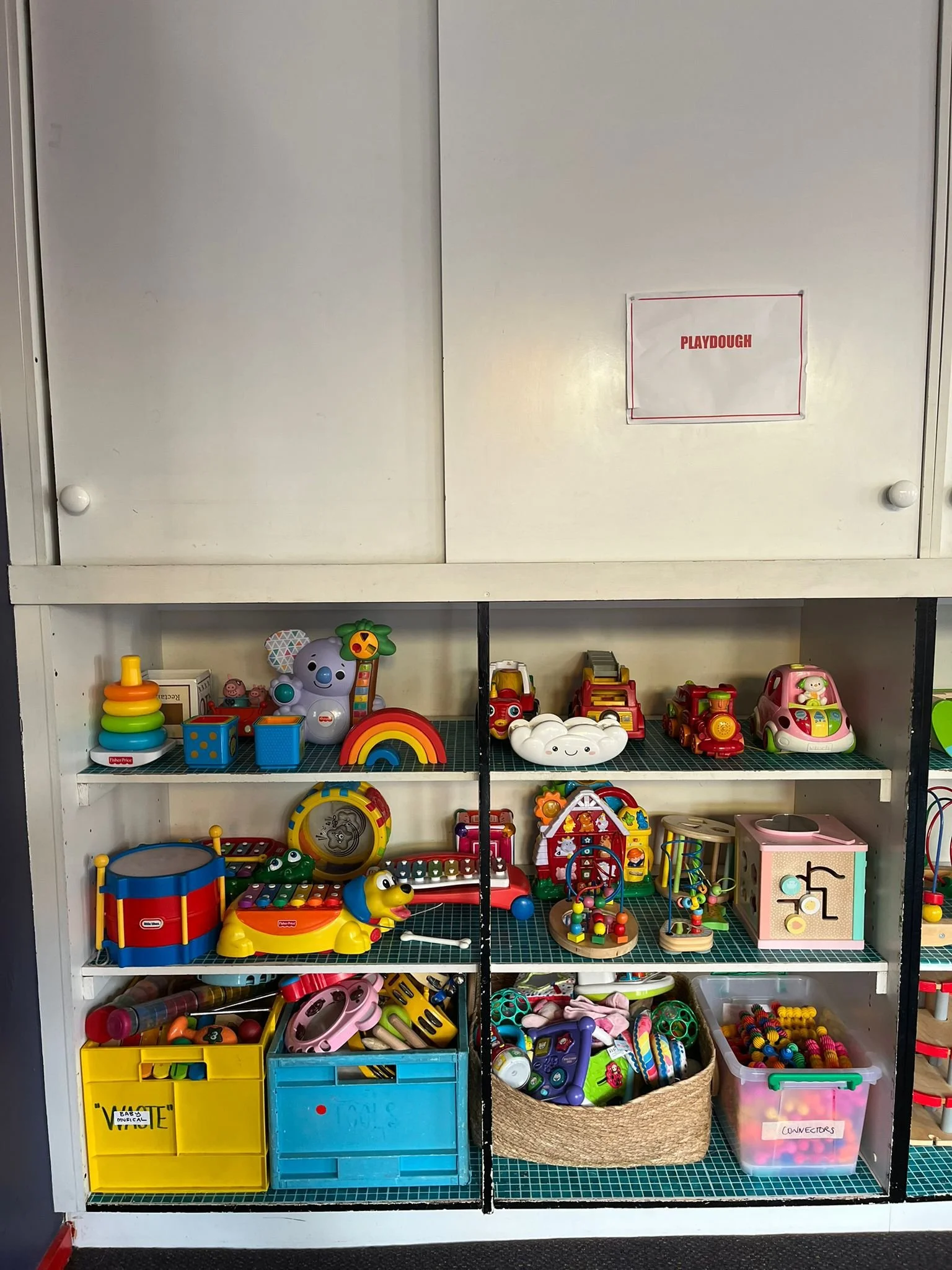 Toy storage shelf filled with colorful children's toys, including stuffed animals, building blocks, toy cars, musical instruments, and activity sets, located in a play area labeled 'Playdough'.