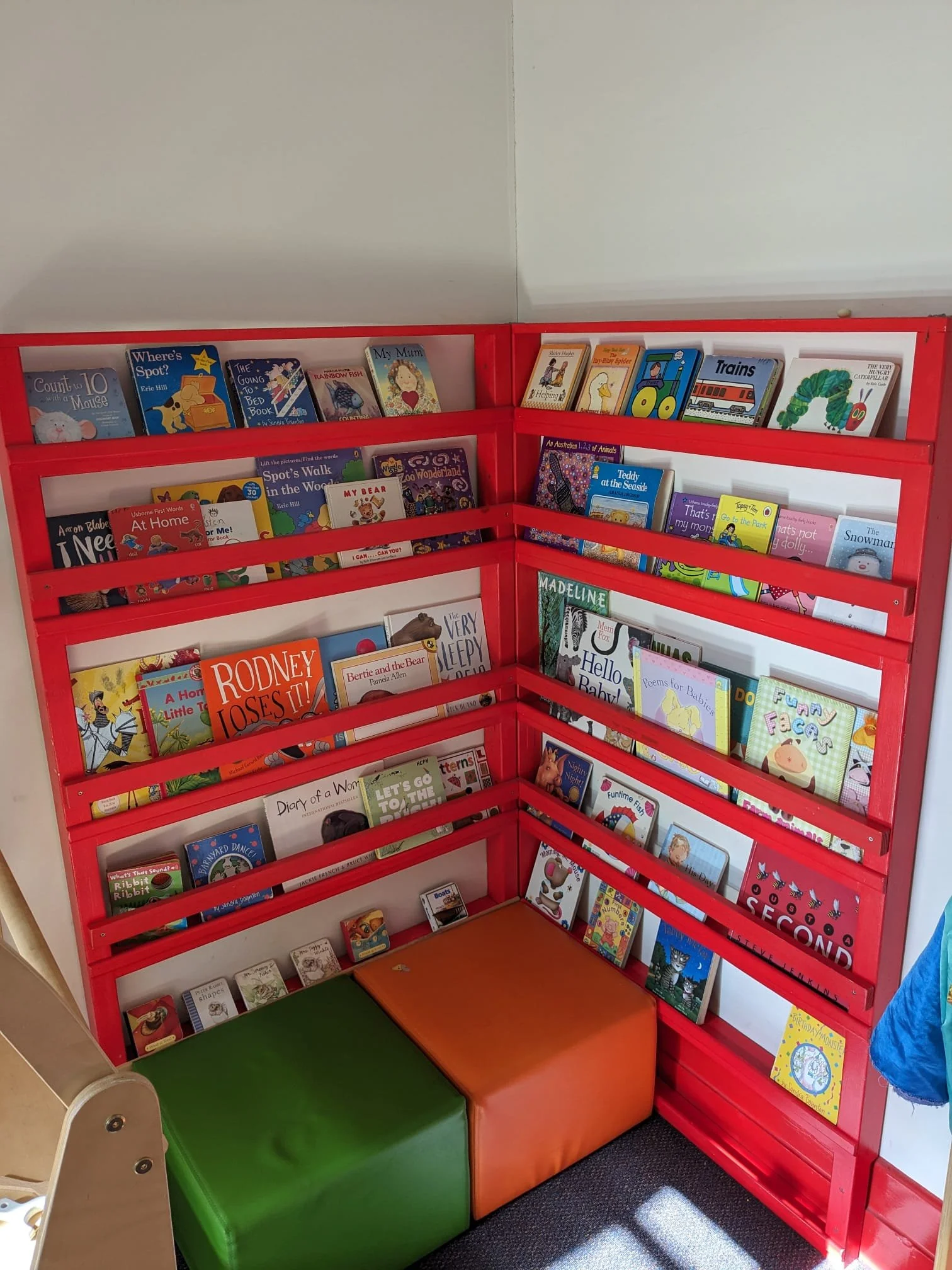 Red bookshelf filled with children's books in a corner, with two colored ottomans in front.