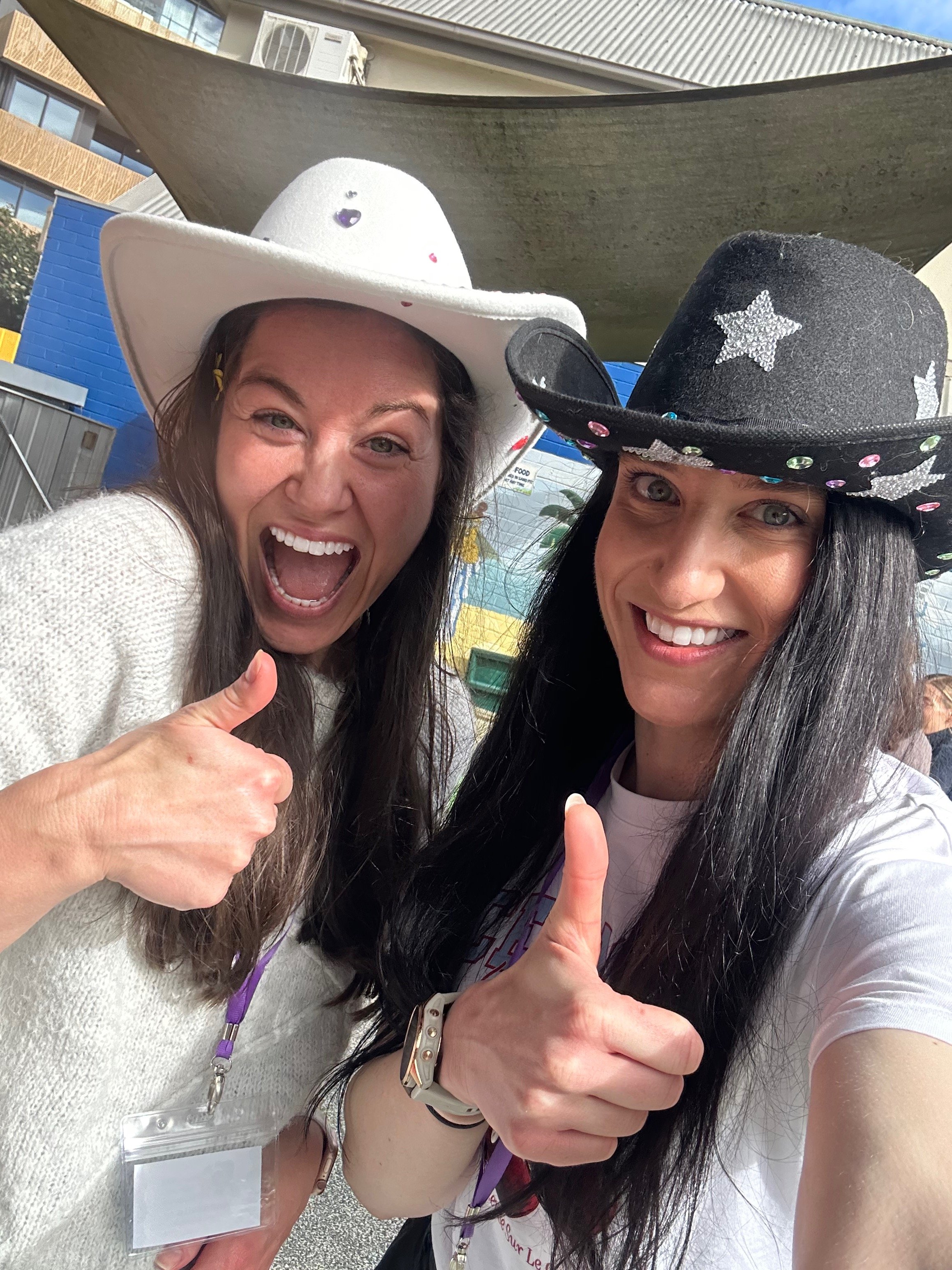 Two women wearing cowboy hats, one white with colorful decorations and one black with silver stars, smiling and giving thumbs up, outdoors under a canopy.