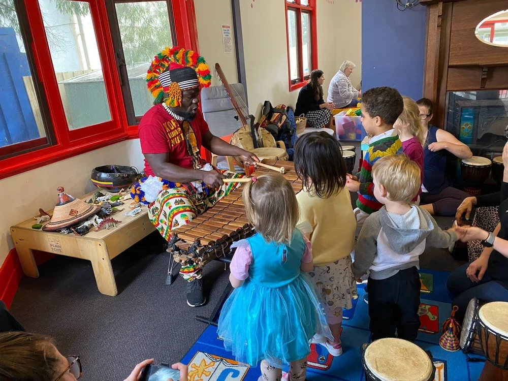 A man in a red shirt and colorful traditional African attire with a headdress demonstrating drum and instrument playing to a group of children in a colorful indoor classroom.
