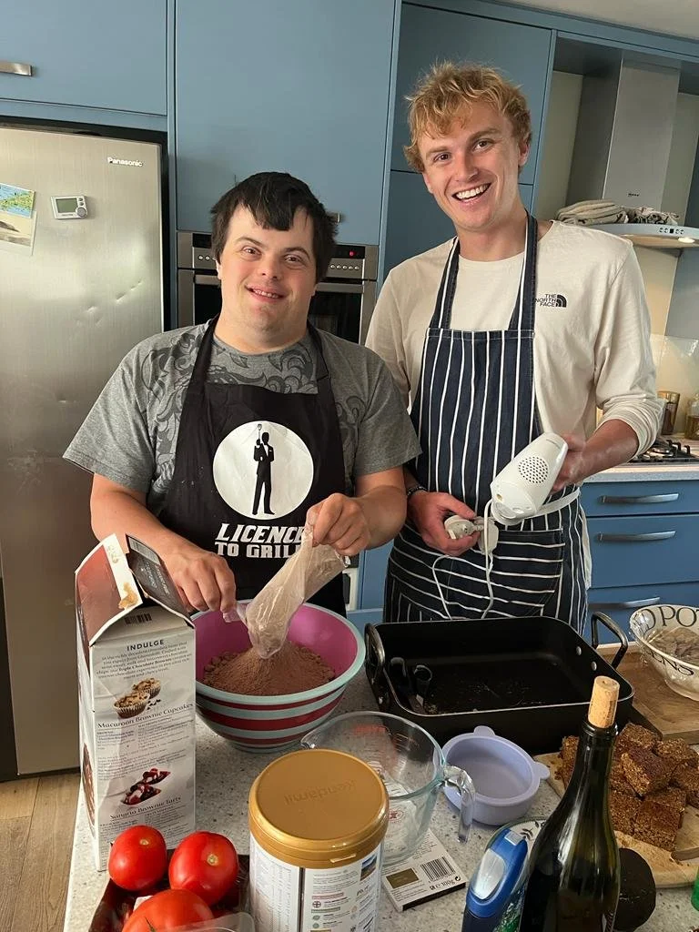 Two men in a kitchen, one with Down syndrome wearing an apron, and a taller man in a striped apron holding a hand mixer, preparing to bake together.