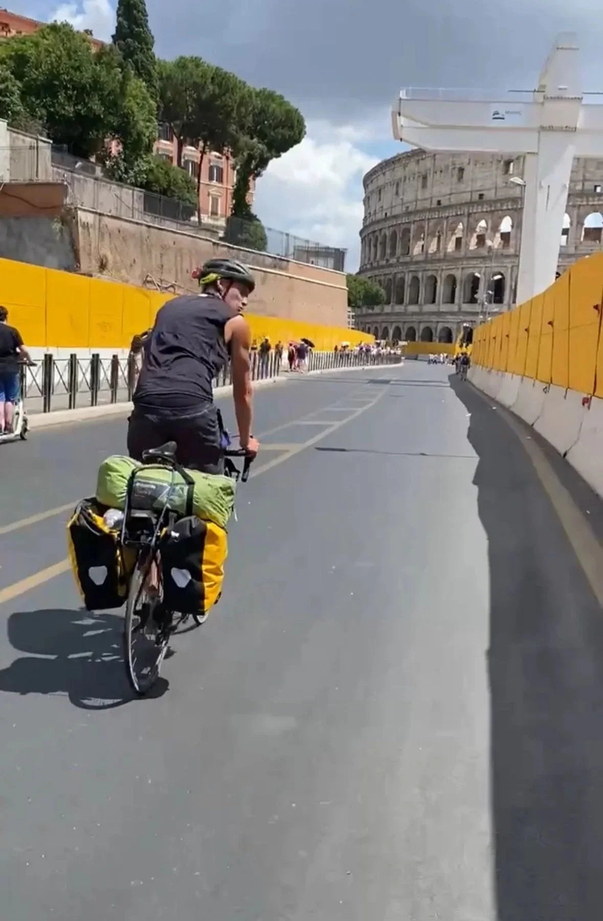 A person riding a bicycle on a road near the Colosseum in Rome, Italy, with yellow barriers lining the sides and a partly cloudy sky overhead.