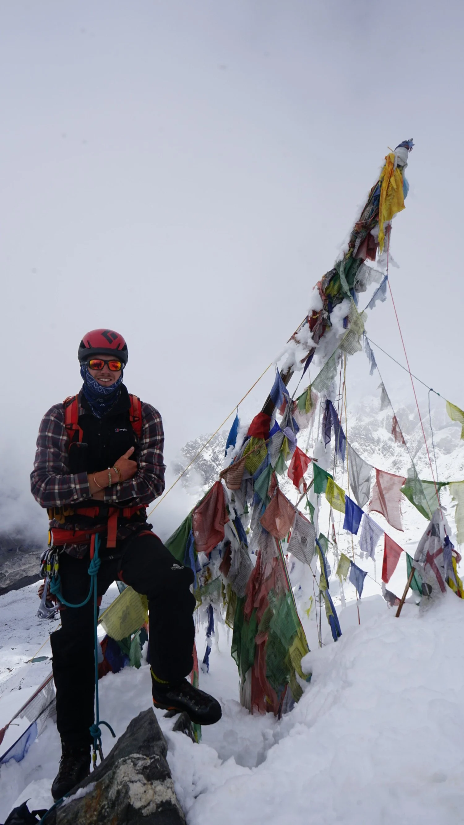 A climber standing next to colorful prayer flags on a snowy mountain, with a foggy background.