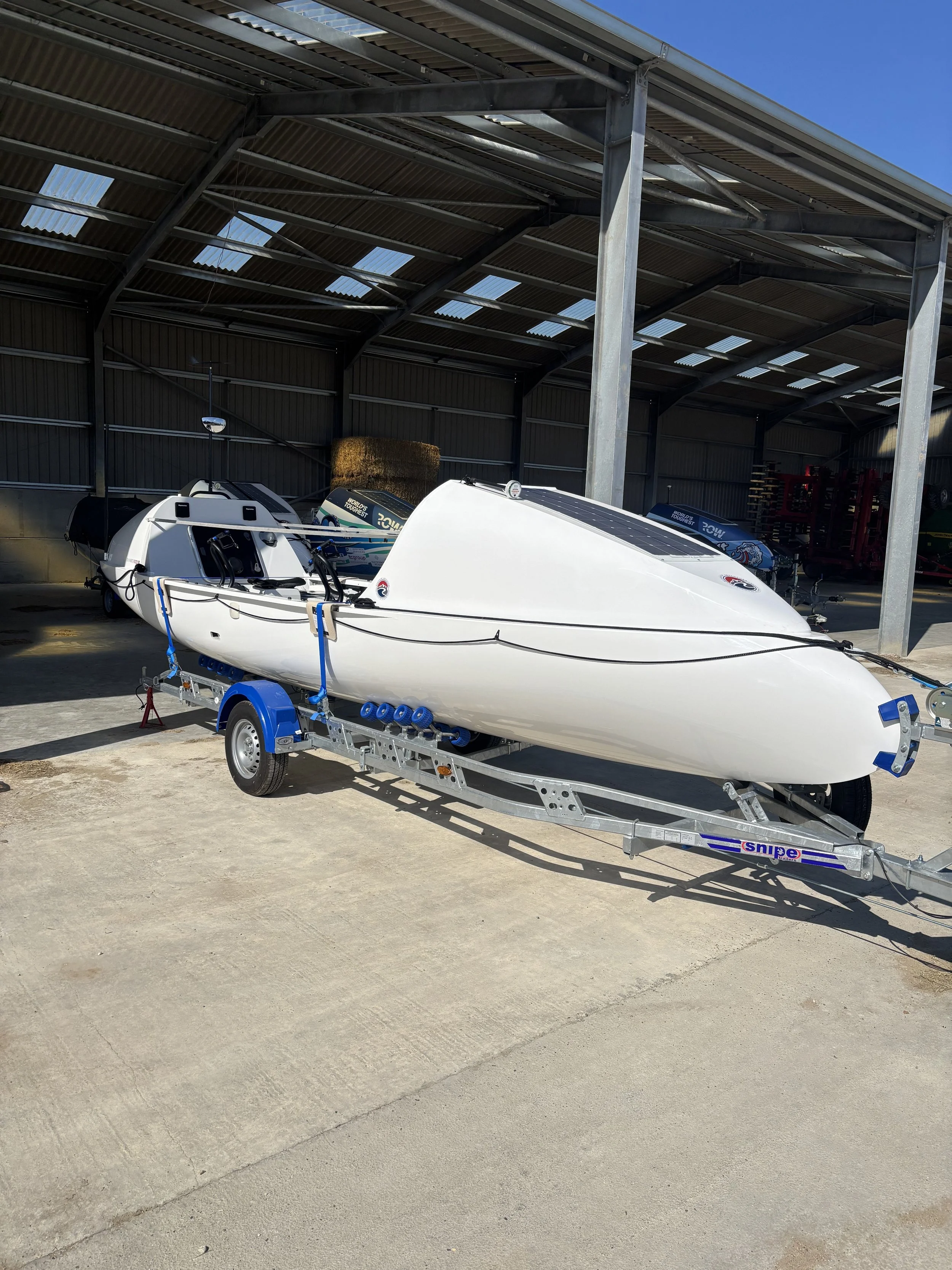 A white racing kayak mounted on a single-axle trailer inside a metal storage shed, with other boats and a hay bale visible in the background.