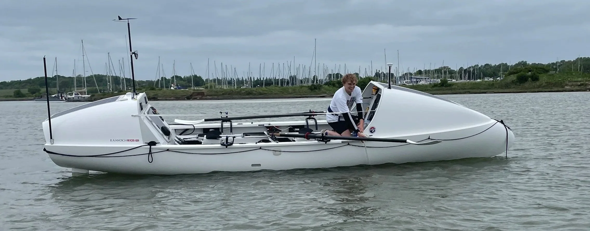 A person on a white racing sailboat with a streamlined hull, preparing to sail on calm water during overcast weather. The boat is equipped with steering and control equipment, and there are multiple sailboats and masts in the background at a marina.