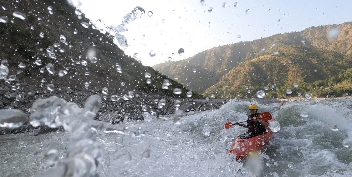 Person kayaking on a river with mountains in the background, water splashing around.