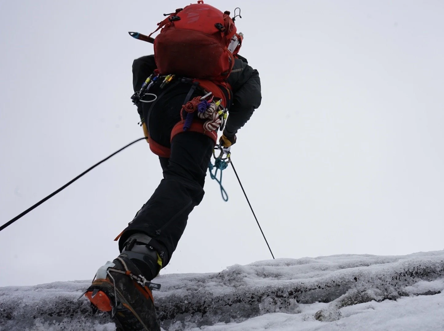 A mountaineer climbing on snow or ice, carrying gear and using safety equipment.