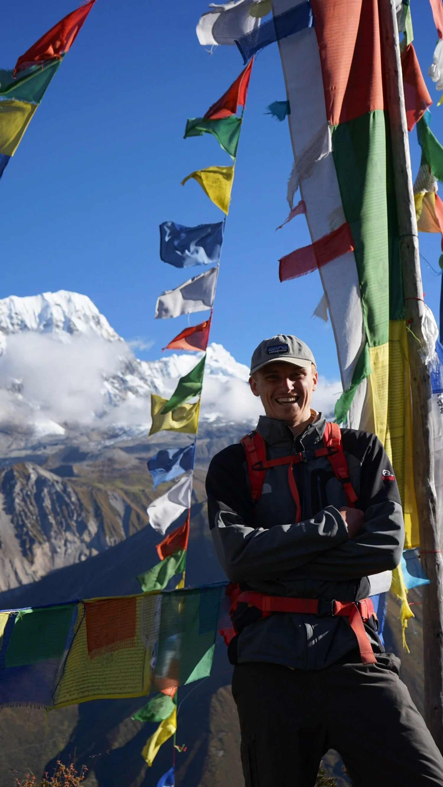 A smiling man standing outdoors in front of colorful prayer flags with mountains and clouds in the background.