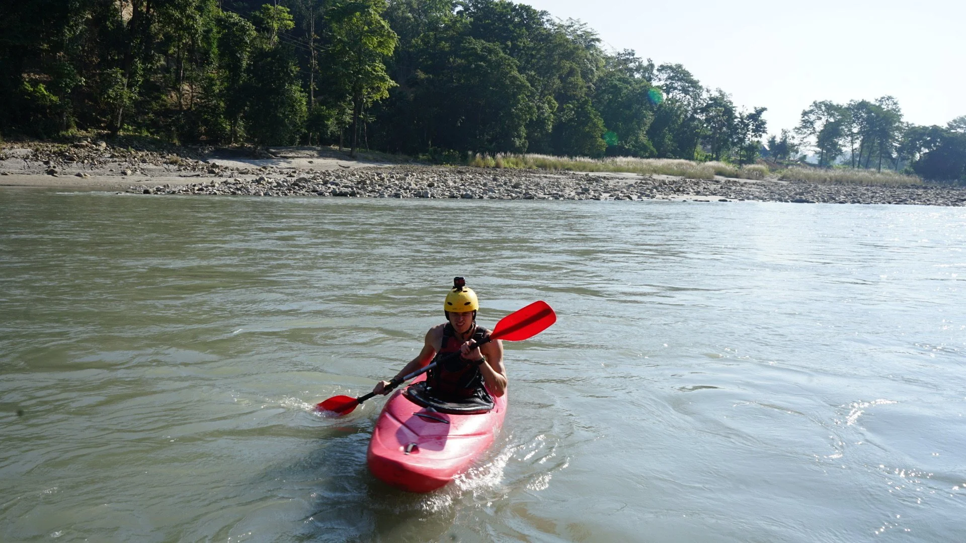 Person kayaking on a river, wearing a yellow helmet and holding a red paddle, with a forested shoreline in the background.
