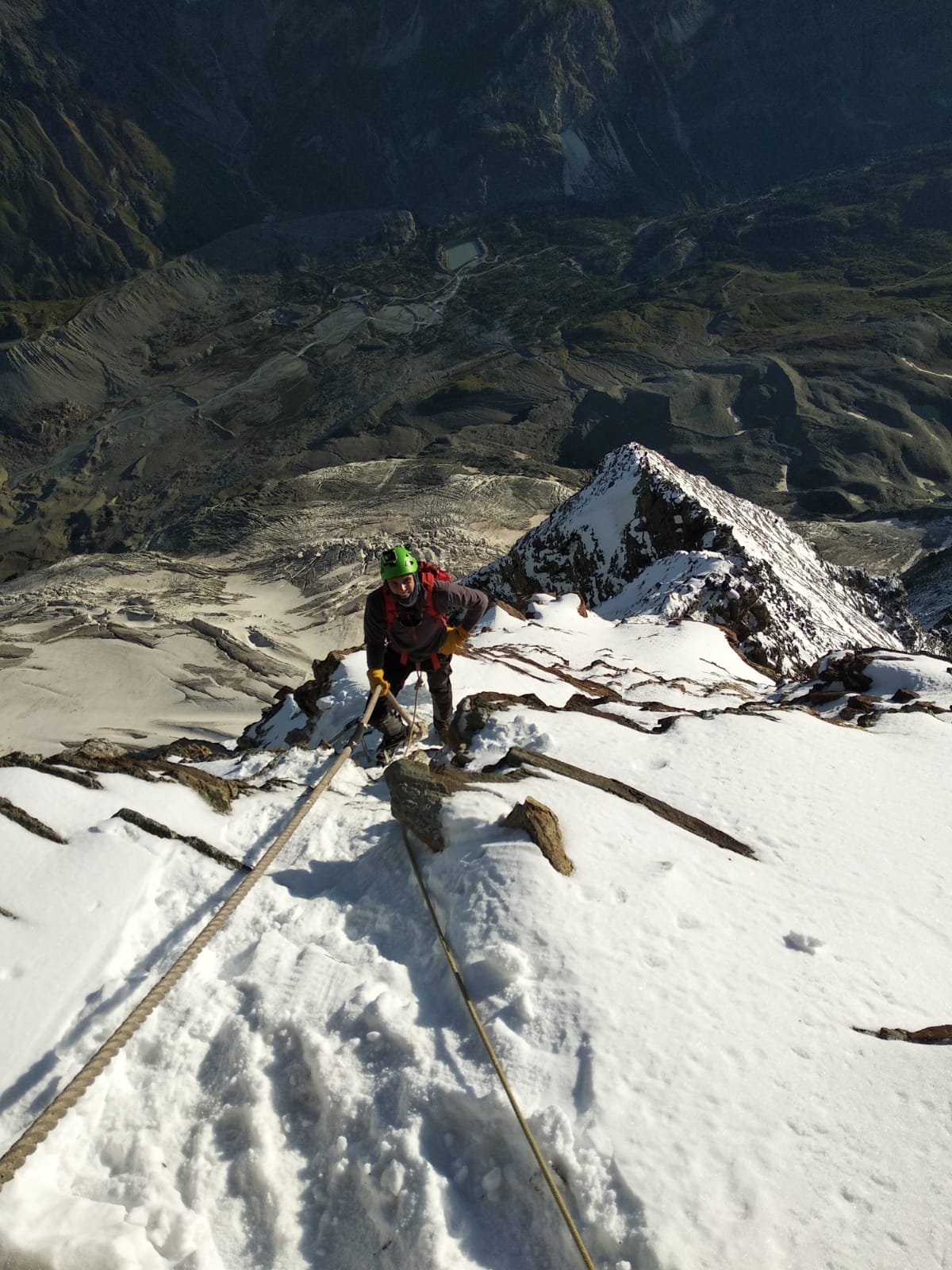 A climber ascending a snowy mountain ridge with a steep drop below, using ropes and safety gear, during daytime with a valley visible in the background.