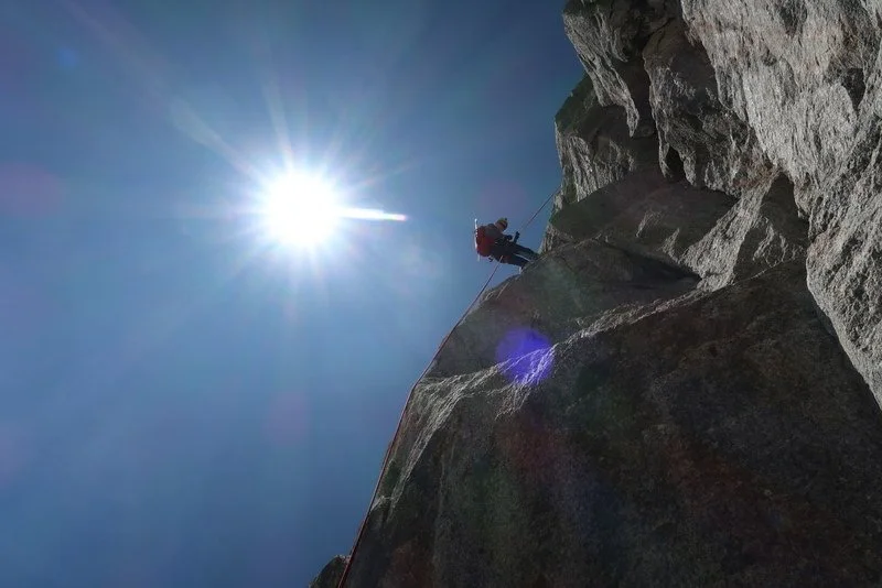 Climber ascending a steep rock face under a bright sun with lens flare.